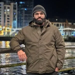 Zain wearing a gray beanie and olive green jacket standing outdoors at night near water with city buildings in the background.