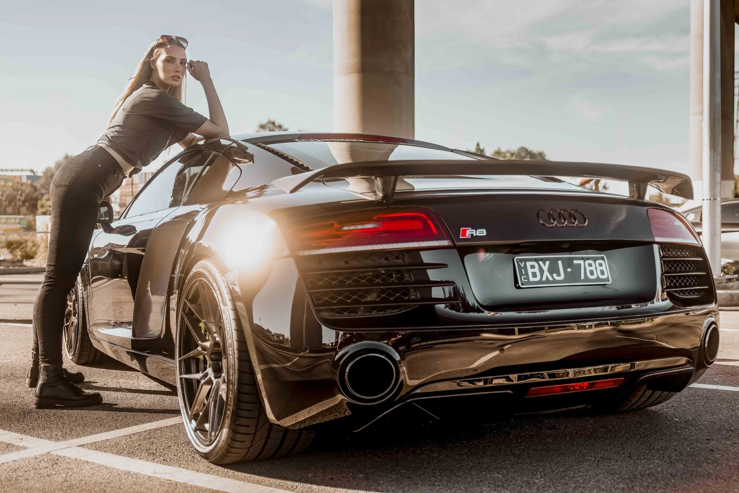 A woman leaning on a black Audi R8 sports car in a parking lot under a bridge, with a sunset or sunrise glow.