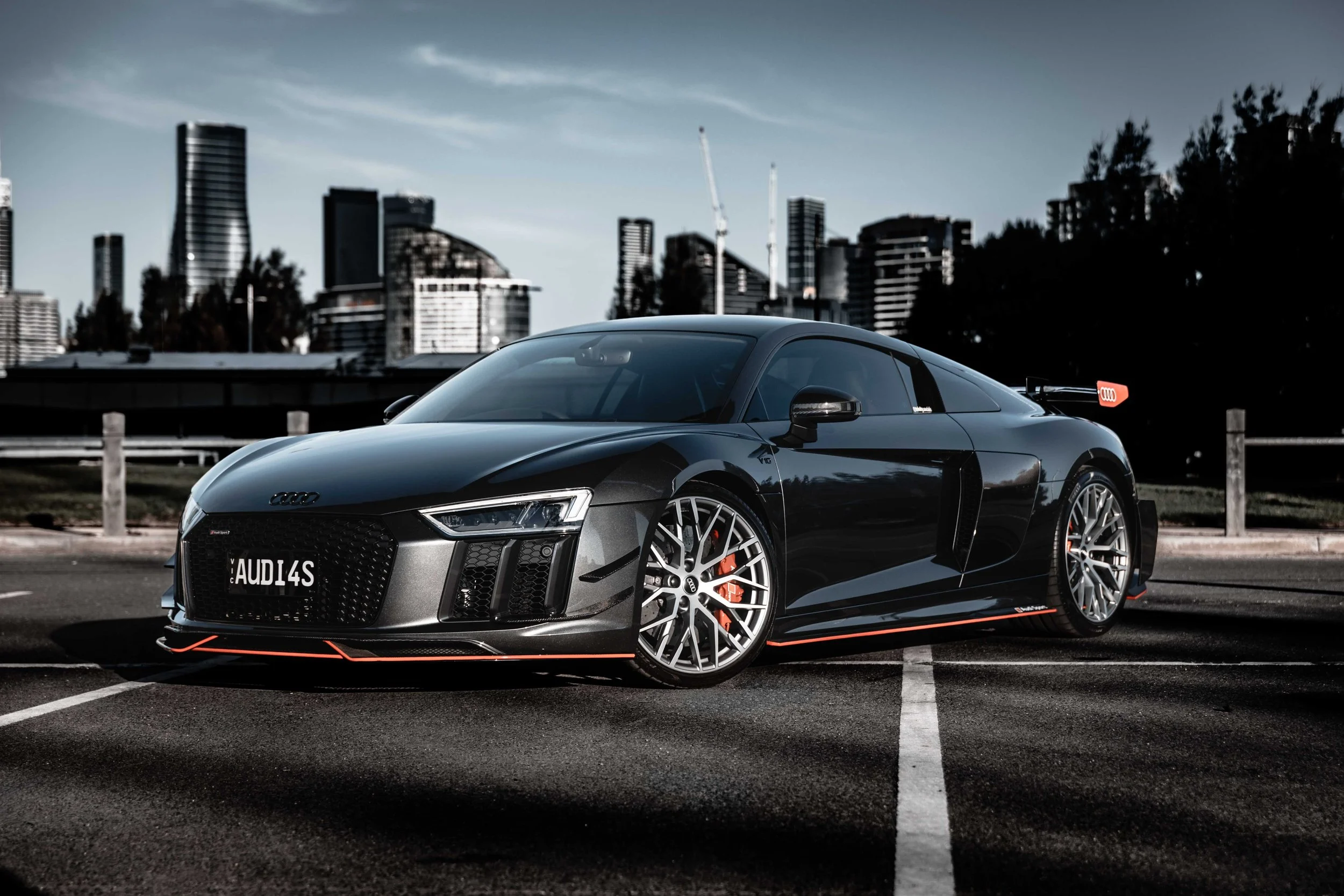 A black Audi sports car parked in an outdoor parking lot with a city skyline featuring tall skyscrapers in the background.