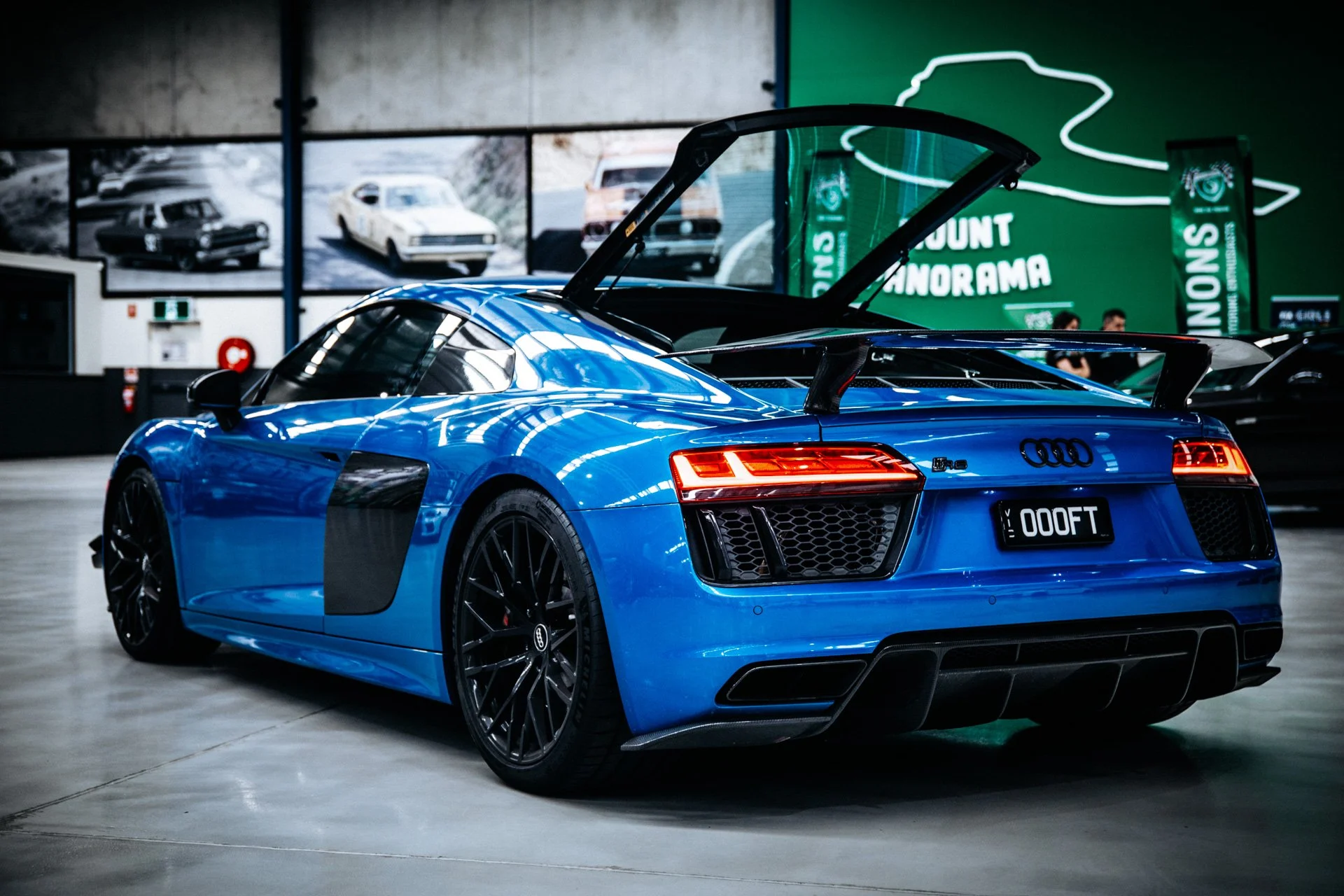 A blue Audi R8 sports car displayed in an indoor showroom, with its rear hatch open, showcasing a sleek design, black wheels, and LED tail lights. The background features posters of vintage cars and a green sign with white text.