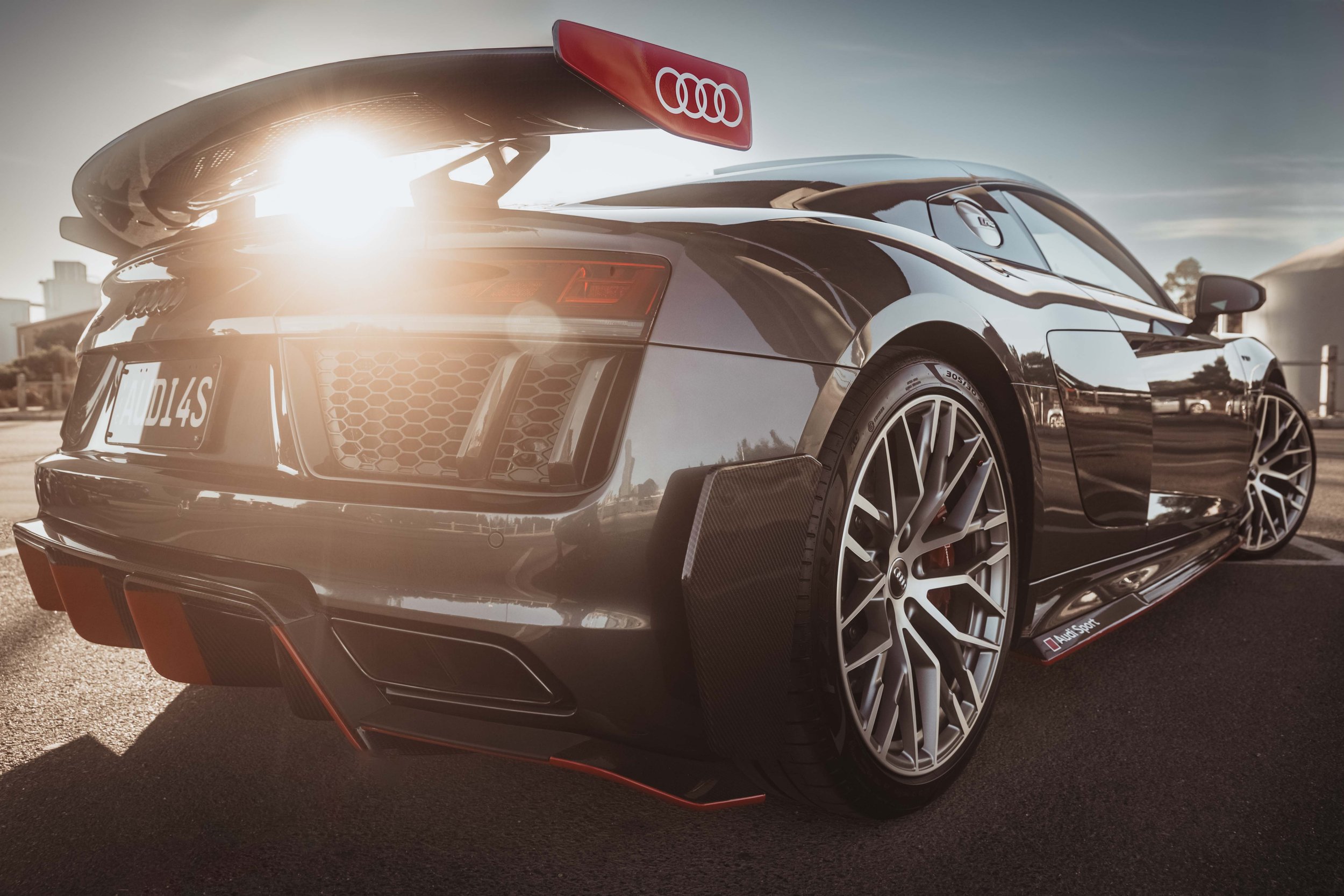 Black Audi sports car with an Audi logo on a red spoiler, parked outdoors in a parking lot during sunset, with reflections on the car's surface and a cityscape in the background.