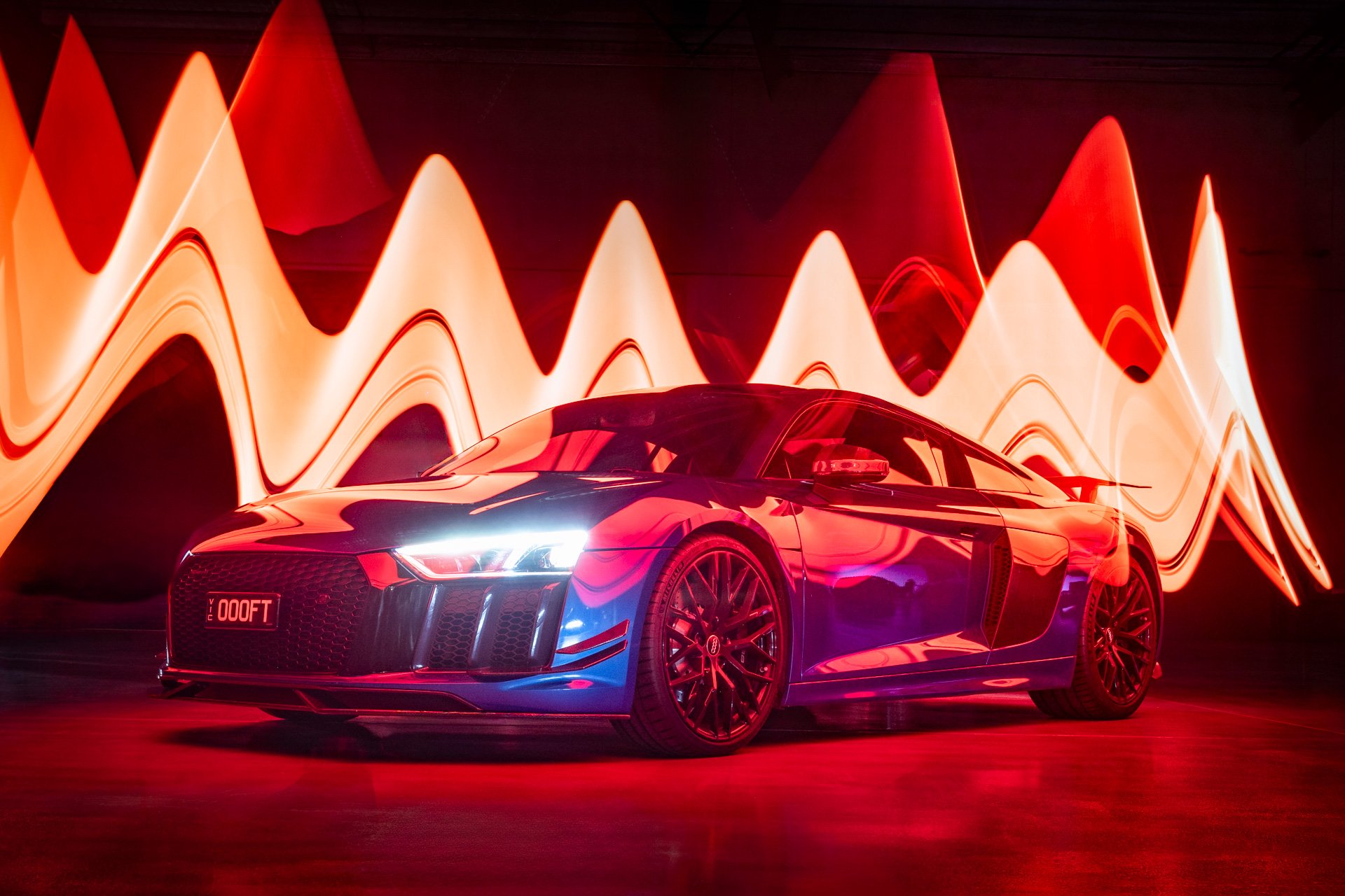 A sleek, black sports car with bright headlamps is illuminated in a room with vibrant, abstract red and orange light patterns on the wall behind it.