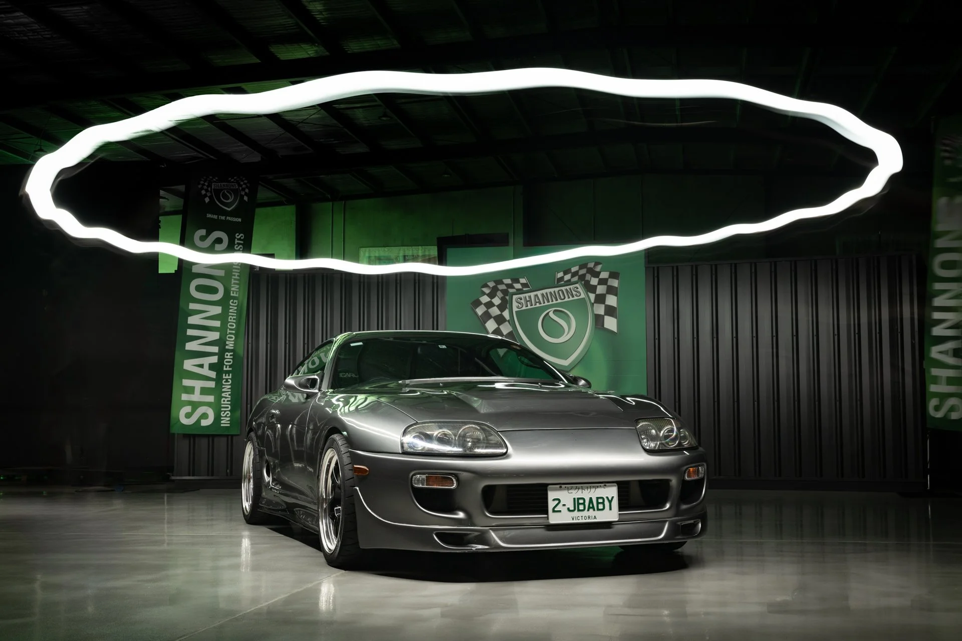 Silver sports car with green license plate that reads '2-JBABY' parked indoors in front of green banners and a black wall, with a large white illuminated light fixture hanging from the ceiling.