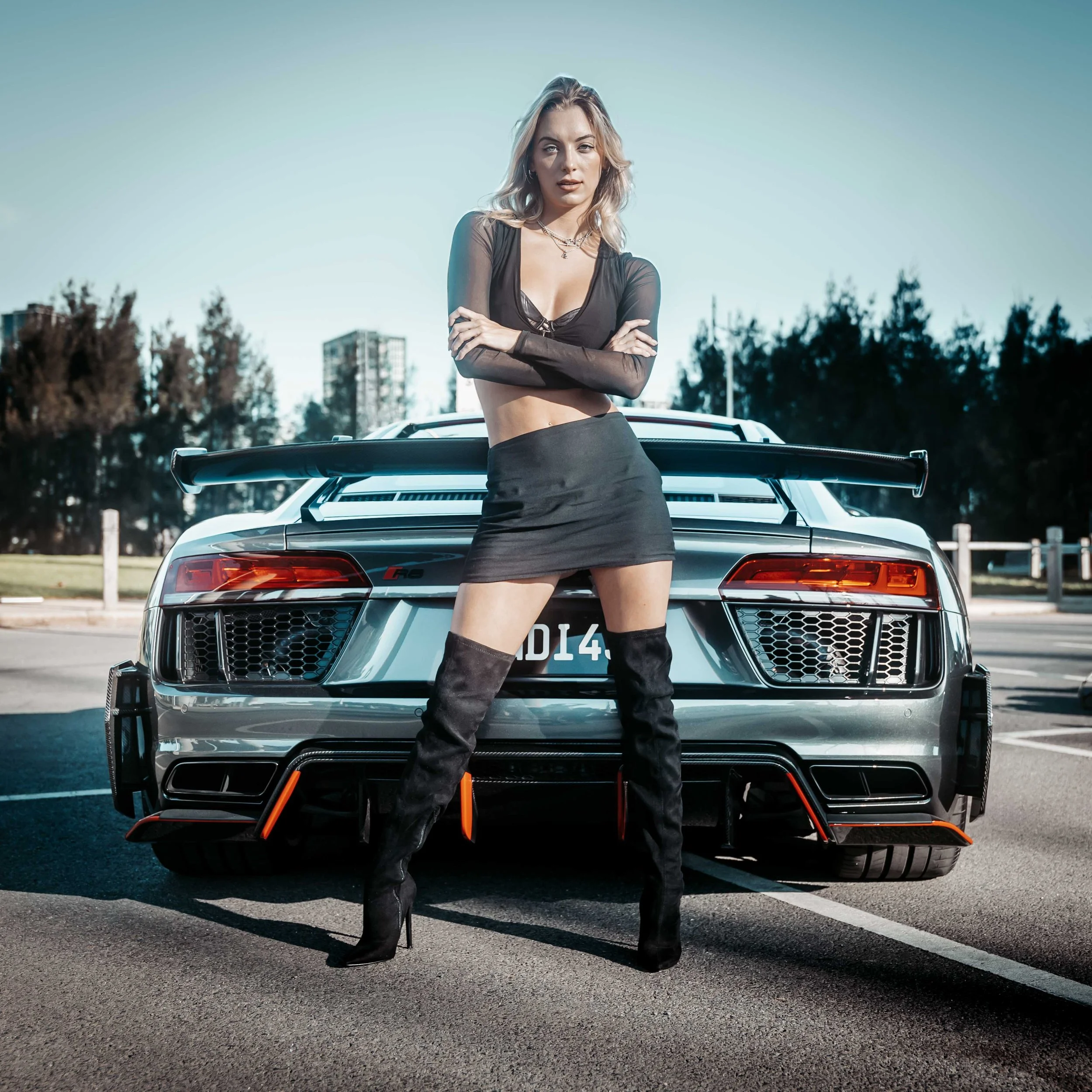 A woman with blonde hair standing in front of a car, wearing a black sheer top, a black skirt, and thigh-high black boots with orange accents, crossing her arms, in an outdoor parking lot with trees and buildings in the background under a clear blue 