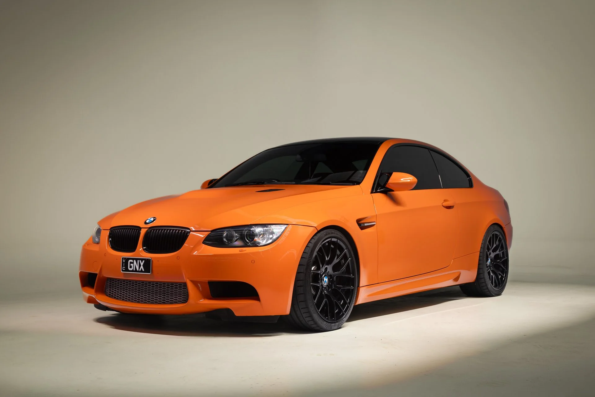 An orange BMW sports car with black wheels and a black roof is displayed against a plain background.