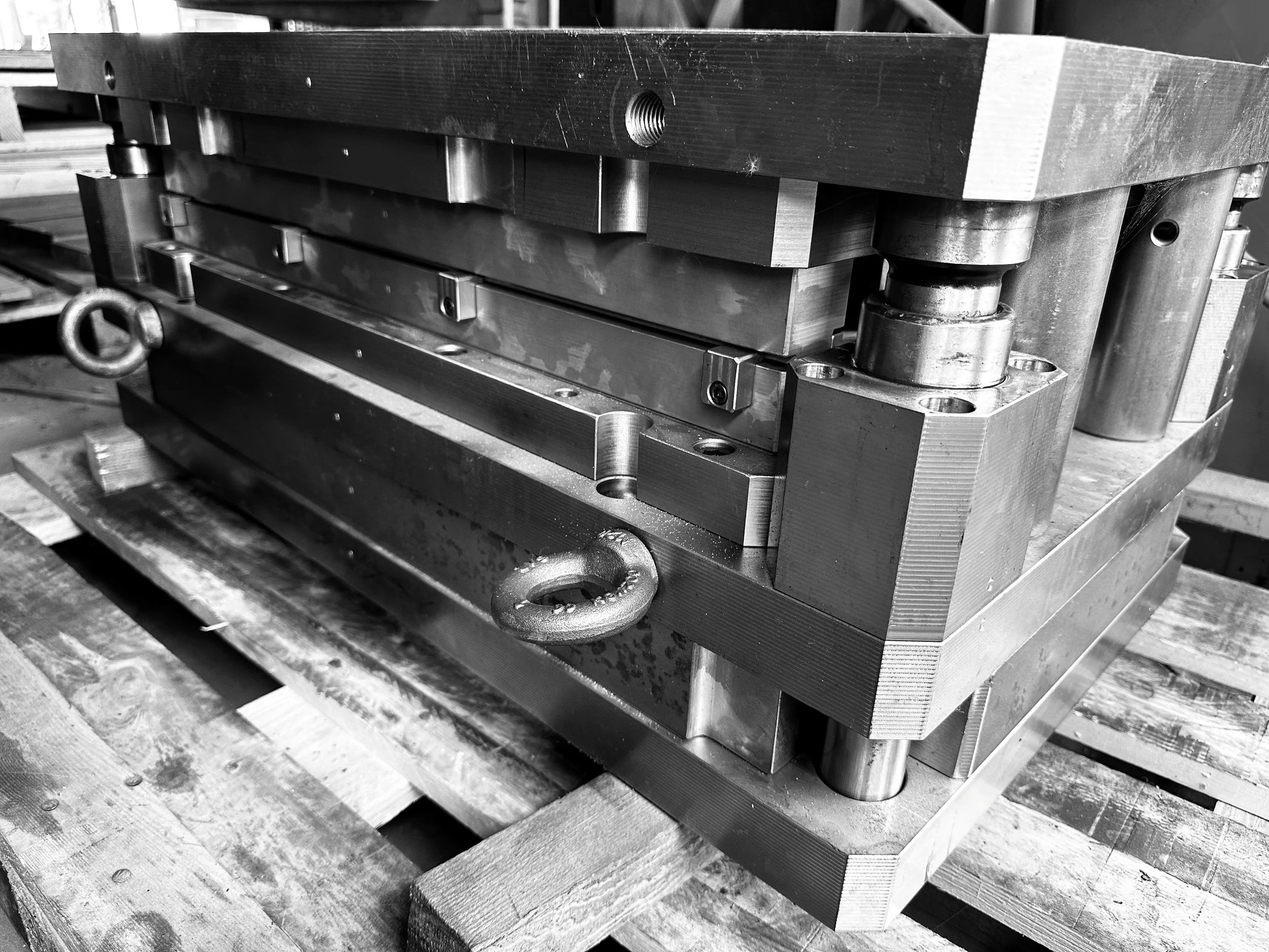 Immagine in bianco e nero di uno stampo meccanico industriale con parti in metallo, verricelli e viti, posizionato su un bancale di legno.