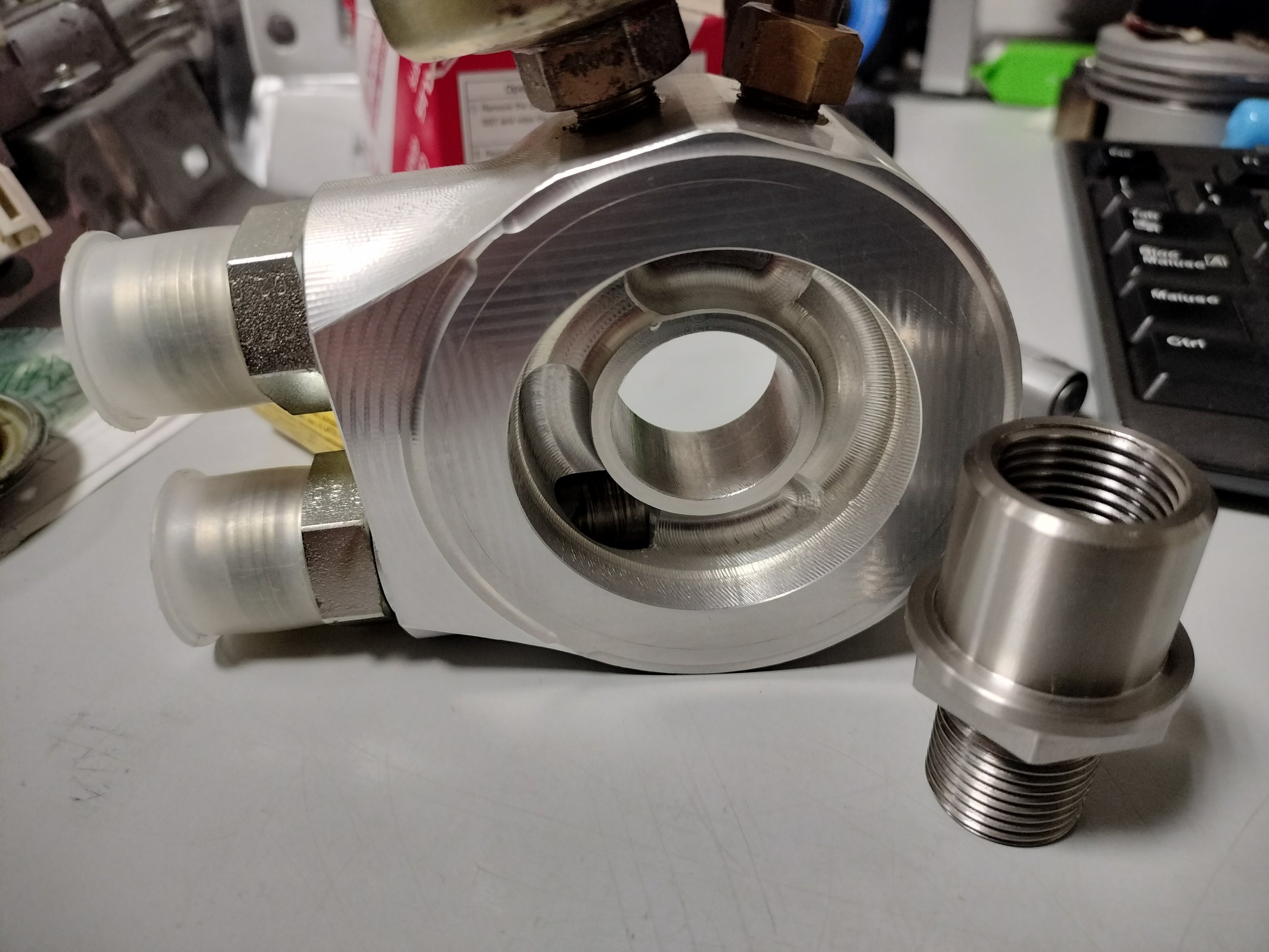 Componente metallico di forma cilindrica con filettature, probabilmente parte di una macchina o un motore, posizionato su una superficie bianca con altri strumenti e oggetti in background.