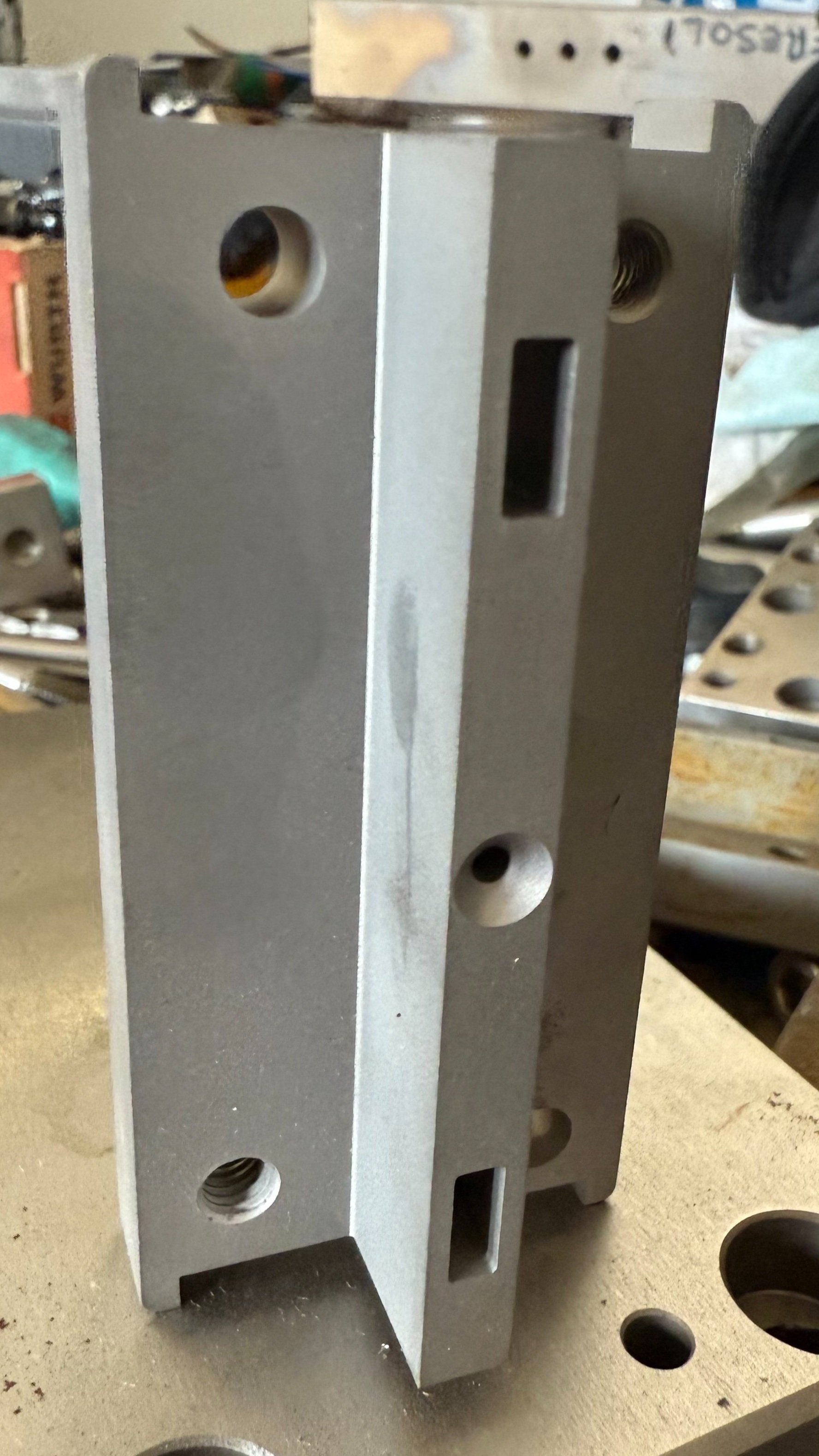 Un componente metallico rettangolare con fori e scanalature, posizionato sopra un banco di lavoro.