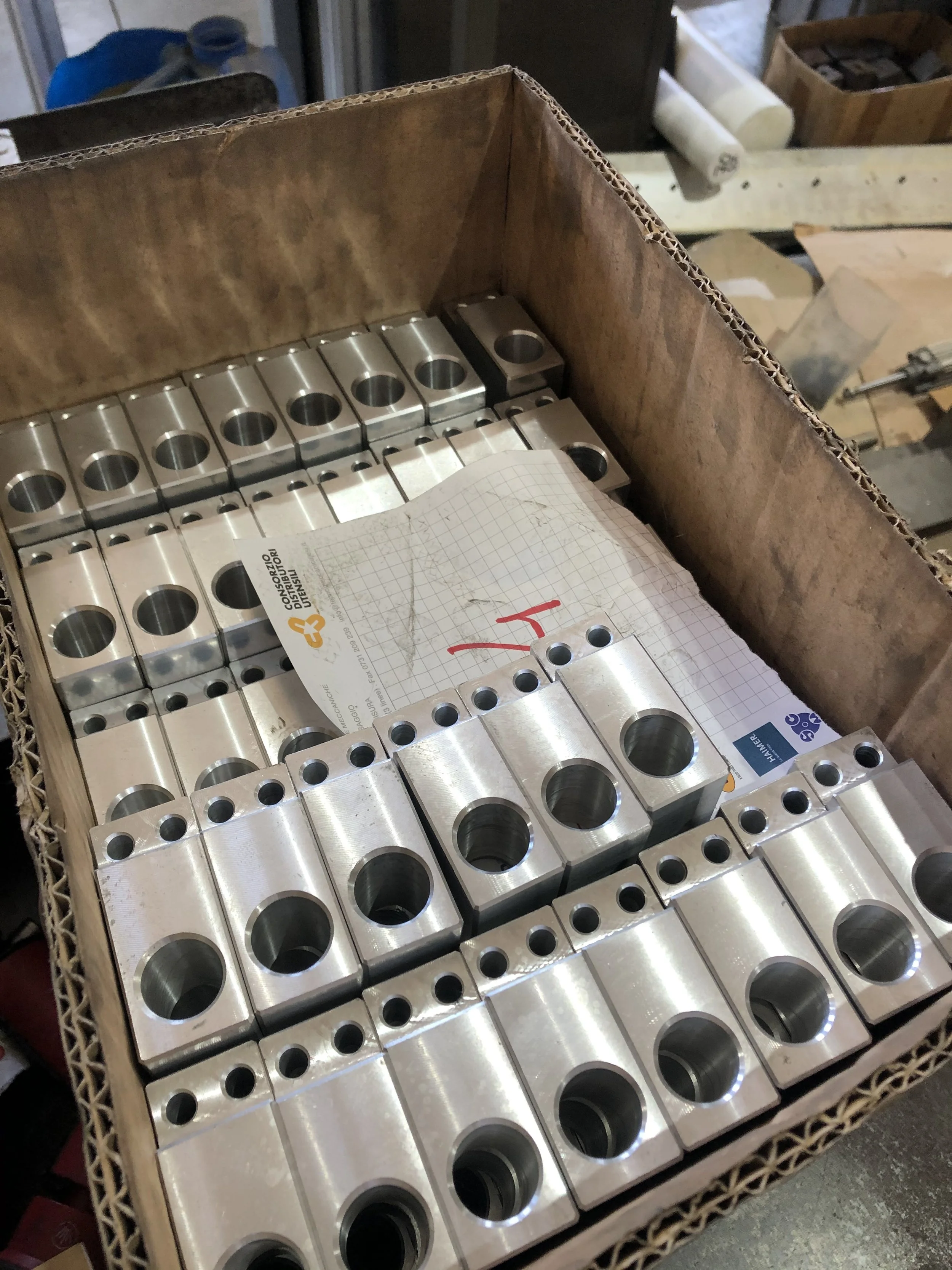 Scatola di blocchi metallici con fori di diverse dimensioni, alcuni con scritte rosse, e carta con grafico e scritte.