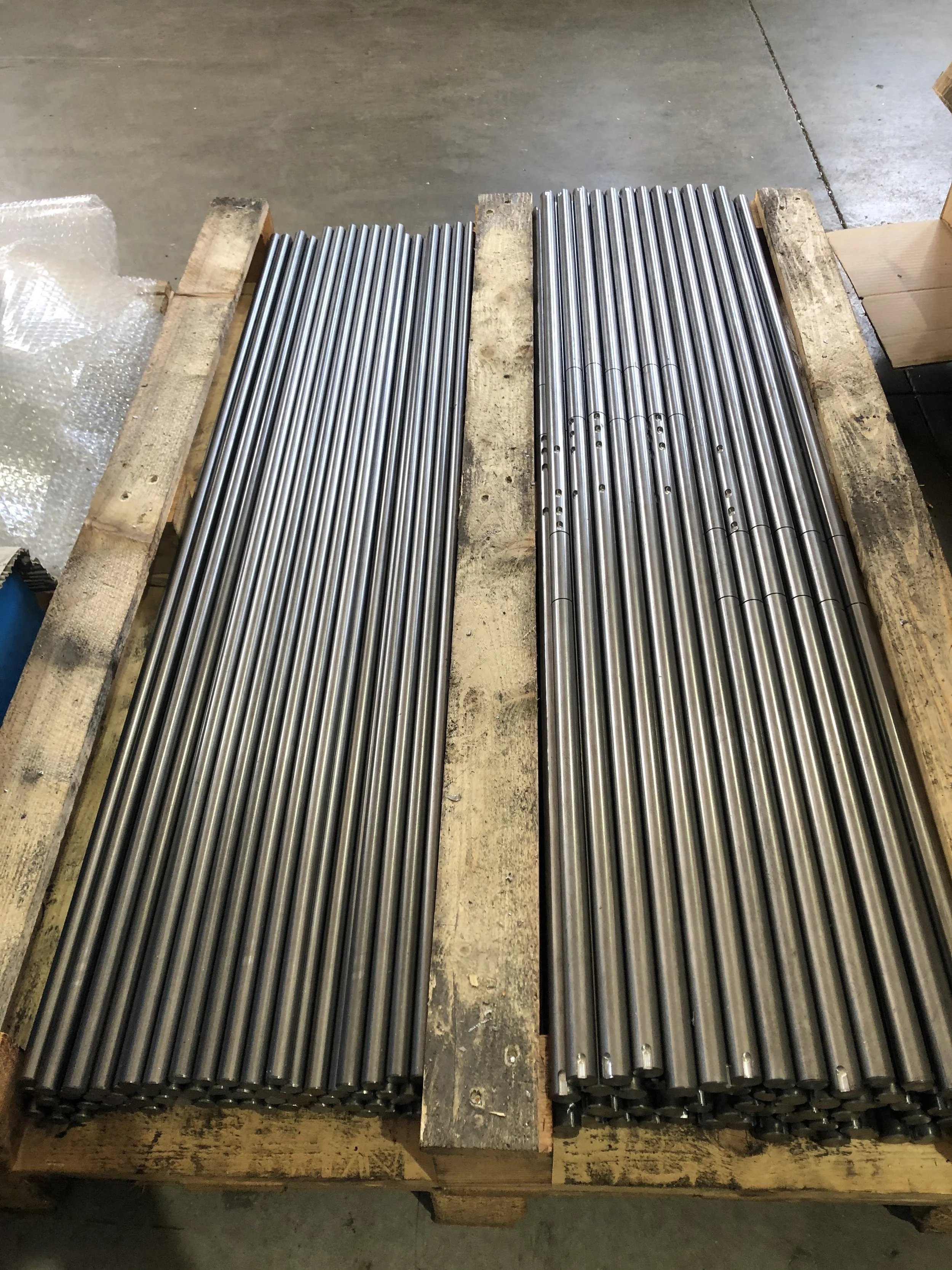 Due gruppi di barre metalliche lunghe e lucide disposte su un pallet di legno, in un ambiente industriale.