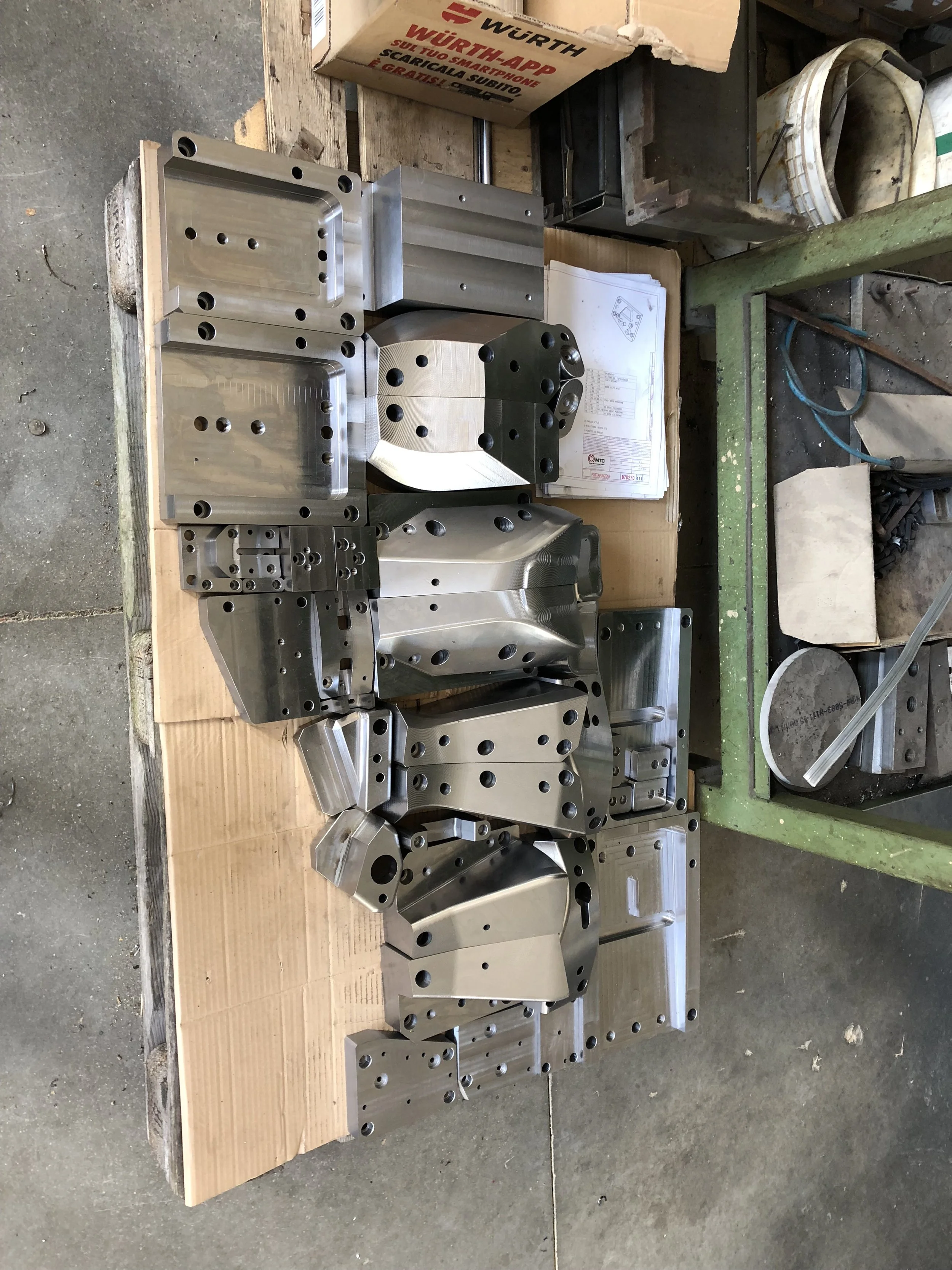 Parti metalliche di macchinari o attrezzature con fori e forme vari, posizionate su un pannello di legno in un'officina