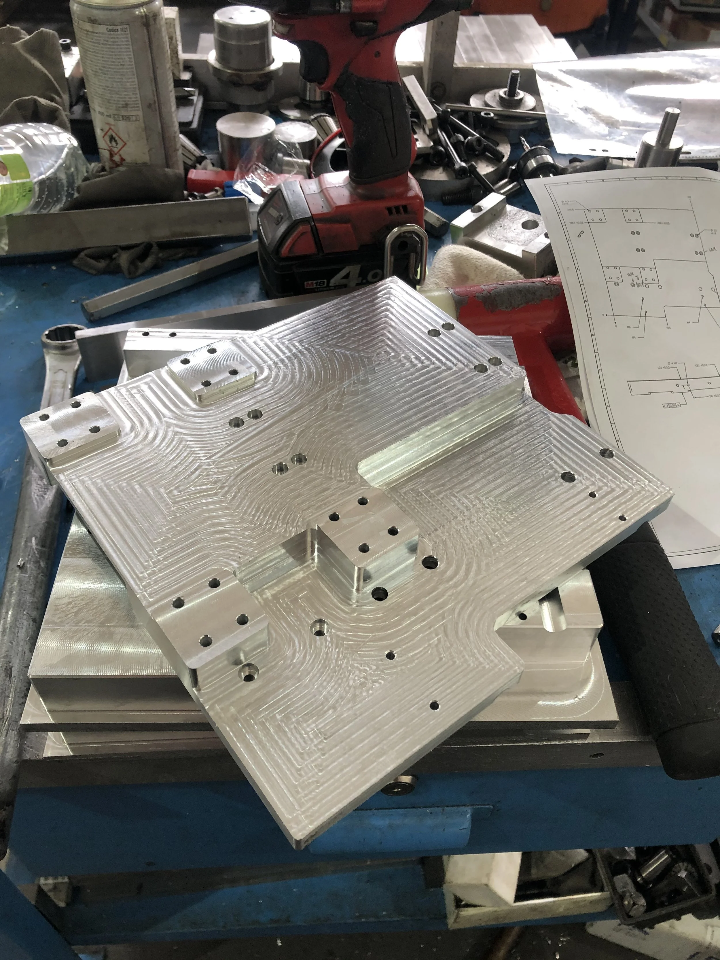 Particolare di un banco da lavoro con un pezzo di metallo lavorato, strumenti, disegni tecnici e una pistola avvitatore.