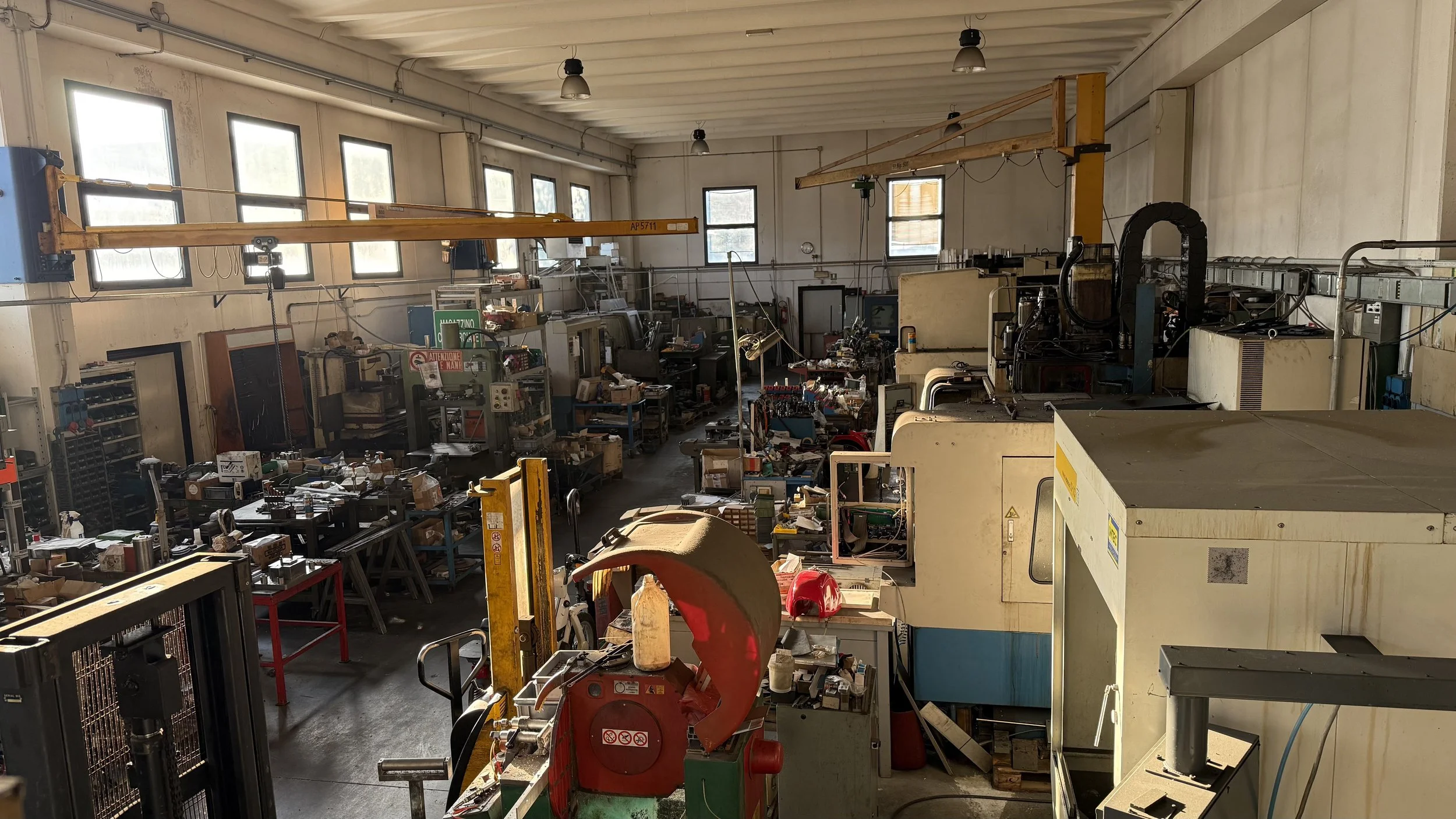 Interno di un'officina con macchinari, attrezzi e scaffali, illuminata dalla luce naturale che entra da diverse finestre.