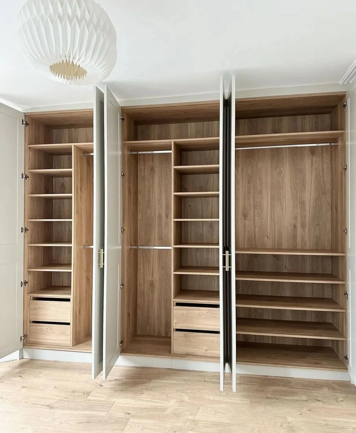 Open wooden closet with three sections, featuring shelves, drawers, and hanging rods, in a room with hardwood flooring and a ceiling light fixture.