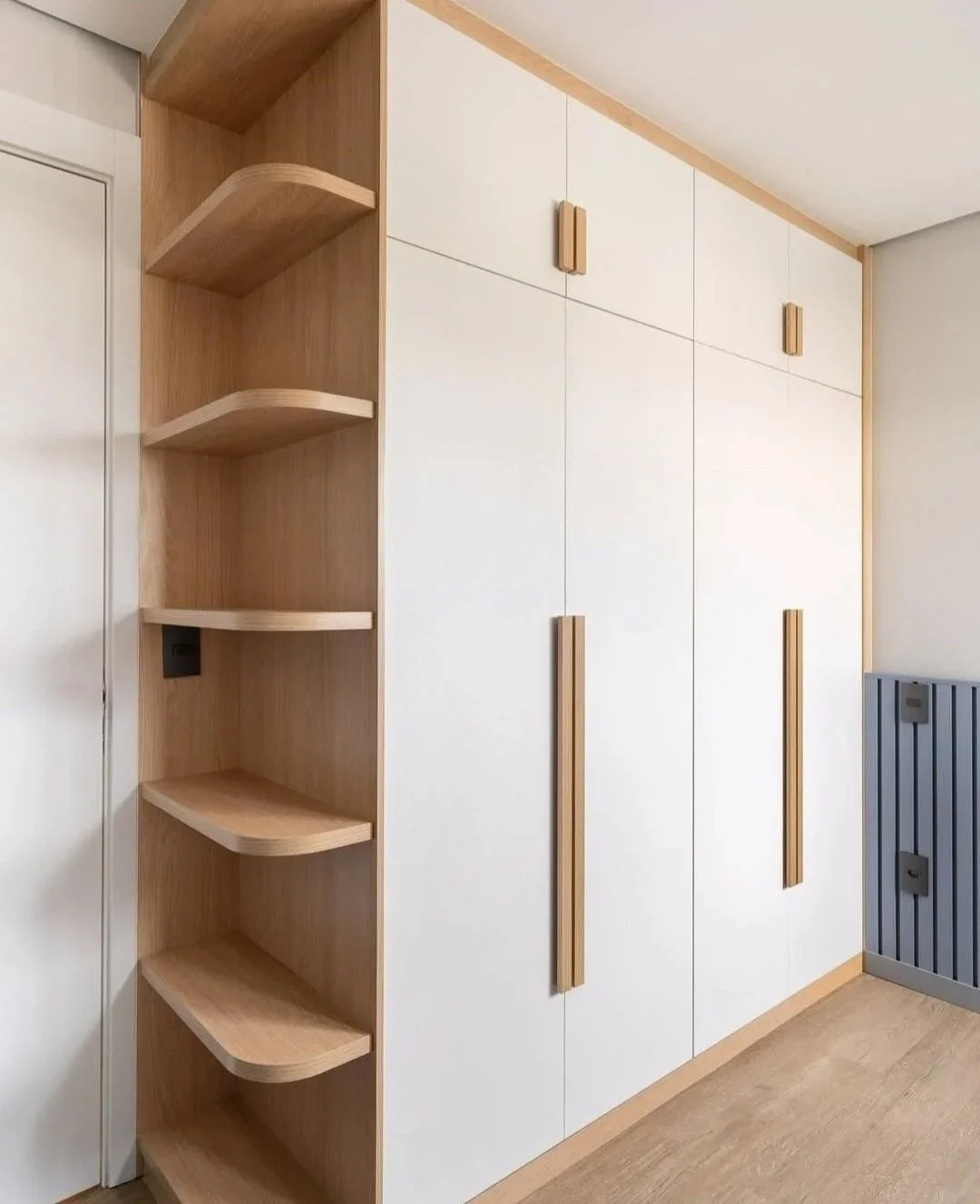 Modern built-in wooden and white cabinet with vertical handles, adjacent to a corner shelving unit, in a room with wood flooring and a gray radiador.