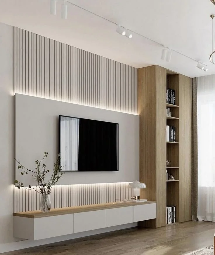 Modern living room with a flat-screen TV on a white wall, a wooden shelf with a vase and books, and a tall wooden bookshelf filled with books, with natural light coming through sheer curtains.