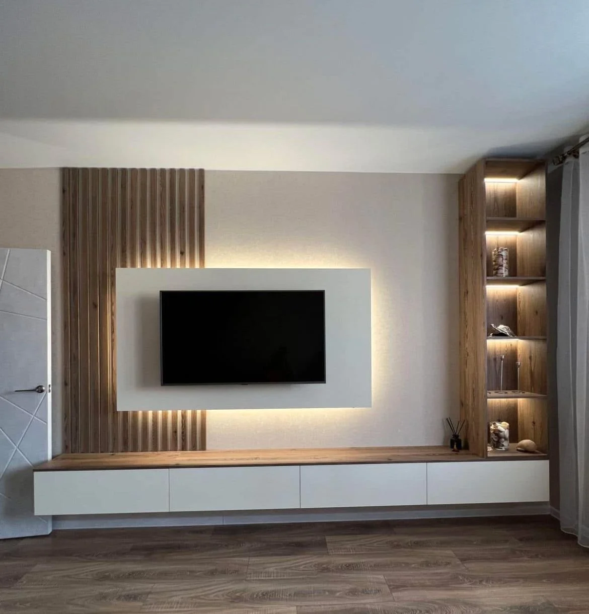 Modern living room with mounted flat-screen TV on a wall with wooden panel background, floating wooden and white TV stand, and built-in wooden bookshelf with decorative items and lighting.