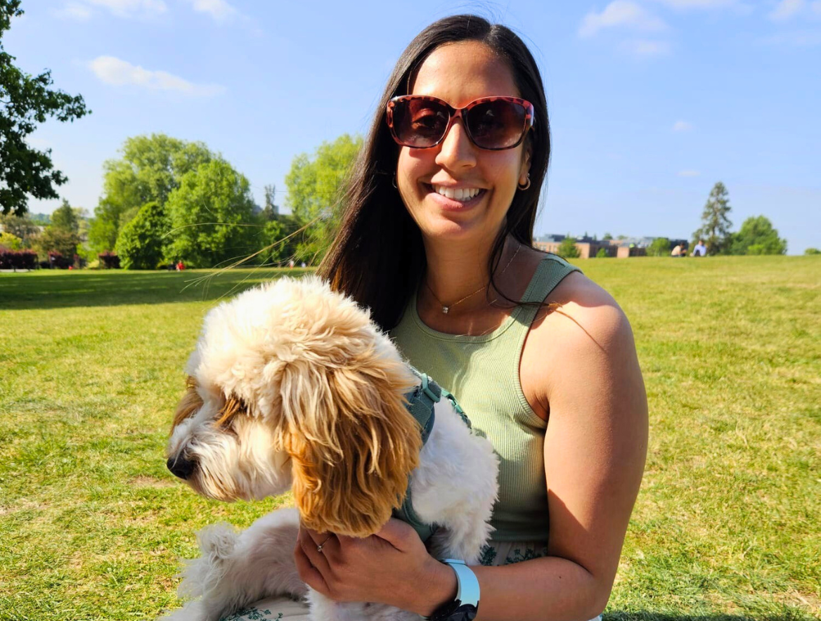 A woman wearing sunglasses and a sleeveless top smiling while holding a fluffy, cream-coloured puppy outdoors on a sunny day with green grass and trees in the background.