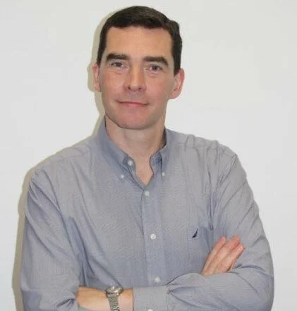 Man with dark hair in a light blue button-up shirt, arms crossed, standing against a plain light background.