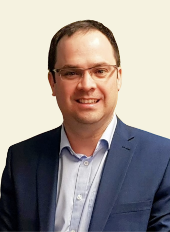 A man wearing glasses, a light blue shirt, and a dark blue suit jacket, smiling against a plain background.