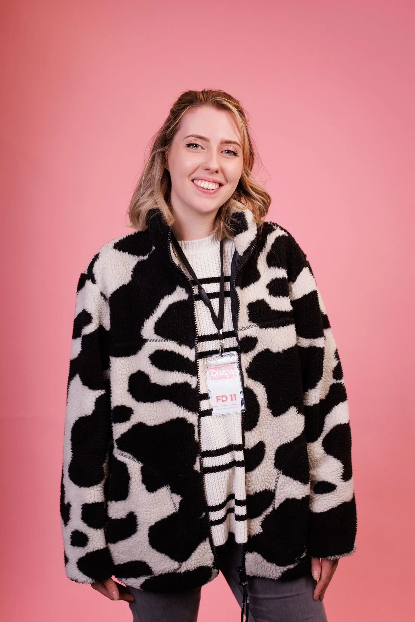 A young woman with shoulder-length blonde hair, smiling, wearing a black and white cow-patterned fleece jacket over a white sweater with black stripes, standing against a pink background.