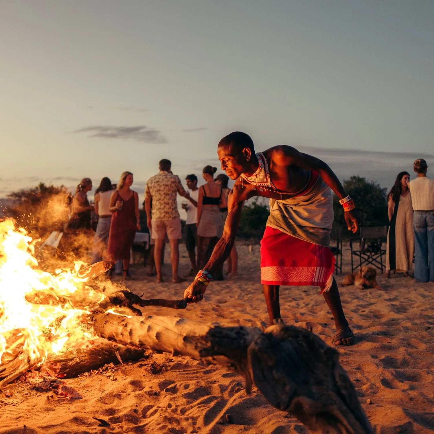 A group of people gathered around a bonfire on a sandy beach during sunset, with one person actively stoking the fire, wearing traditional clothing and jewelry, with others socializing in the background.