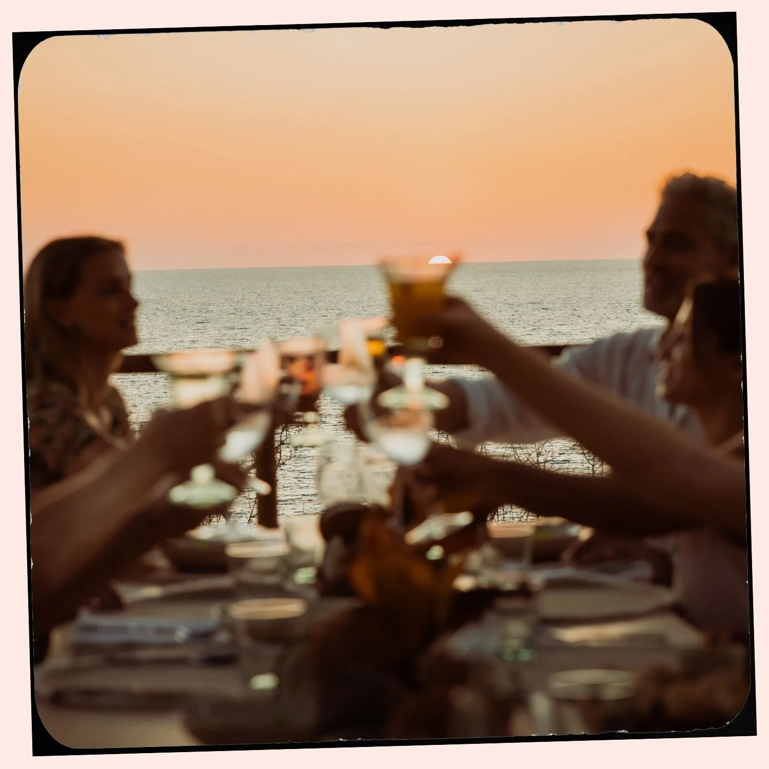 A group of people enjoying a dinner or celebration on a patio by the ocean during sunset, raising glasses in a toast.