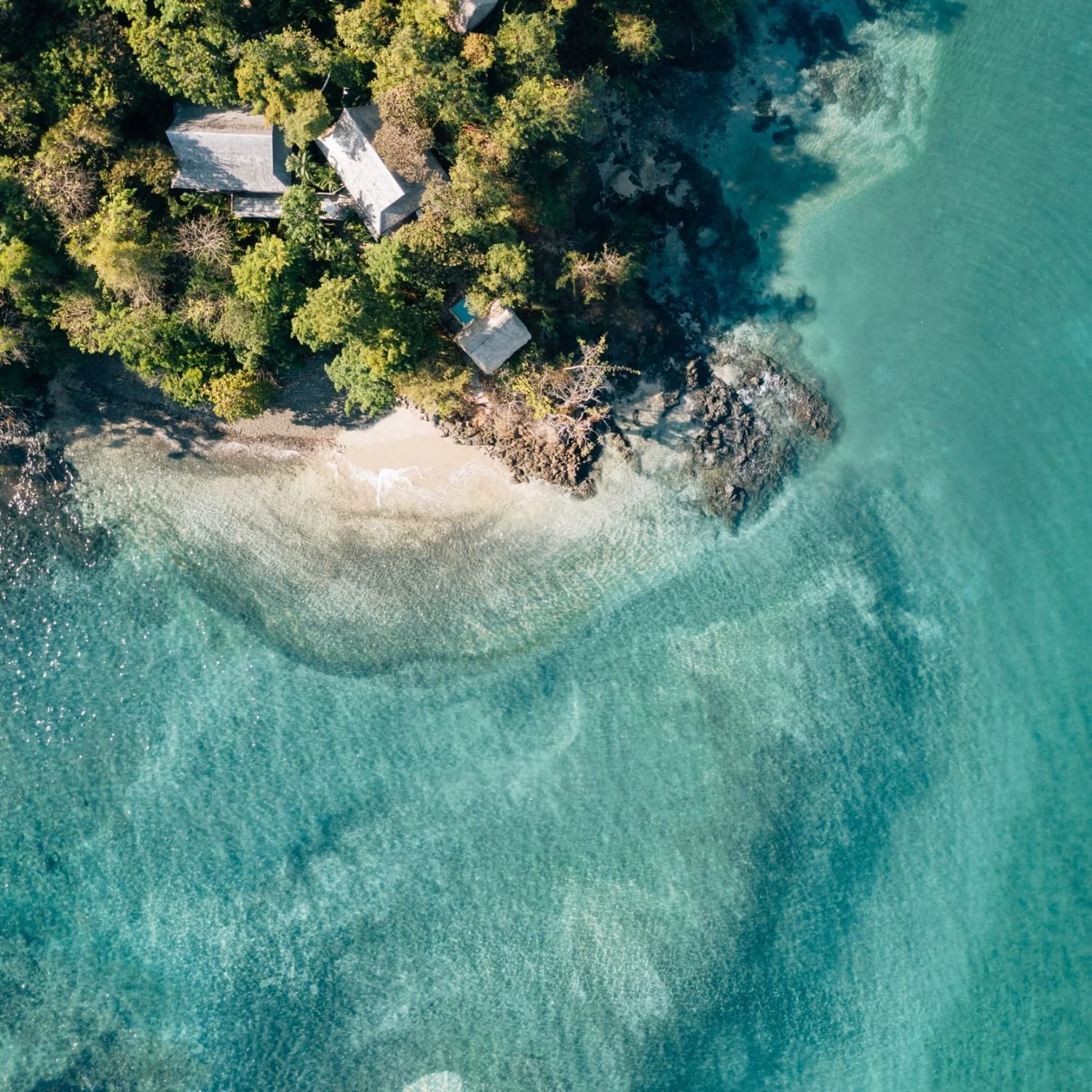 An aerial view of a rocky beach with sand, surrounded by dense green trees and clear turquoise water.