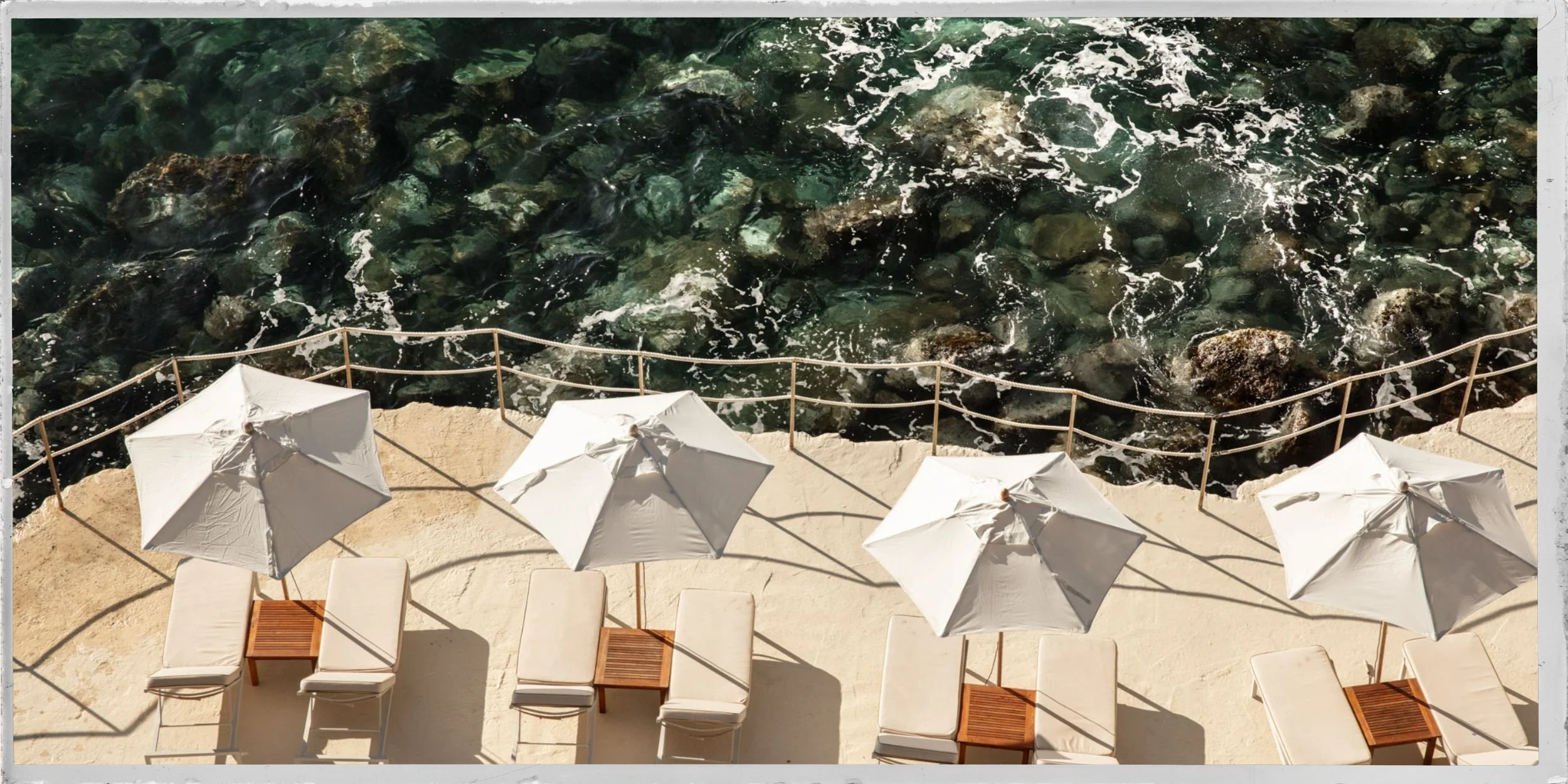 Aerial view of a seaside lounge area with four white sun umbrellas, beige lounge chairs, small wooden tables, and a guardrail, overlooking rocky water with waves.