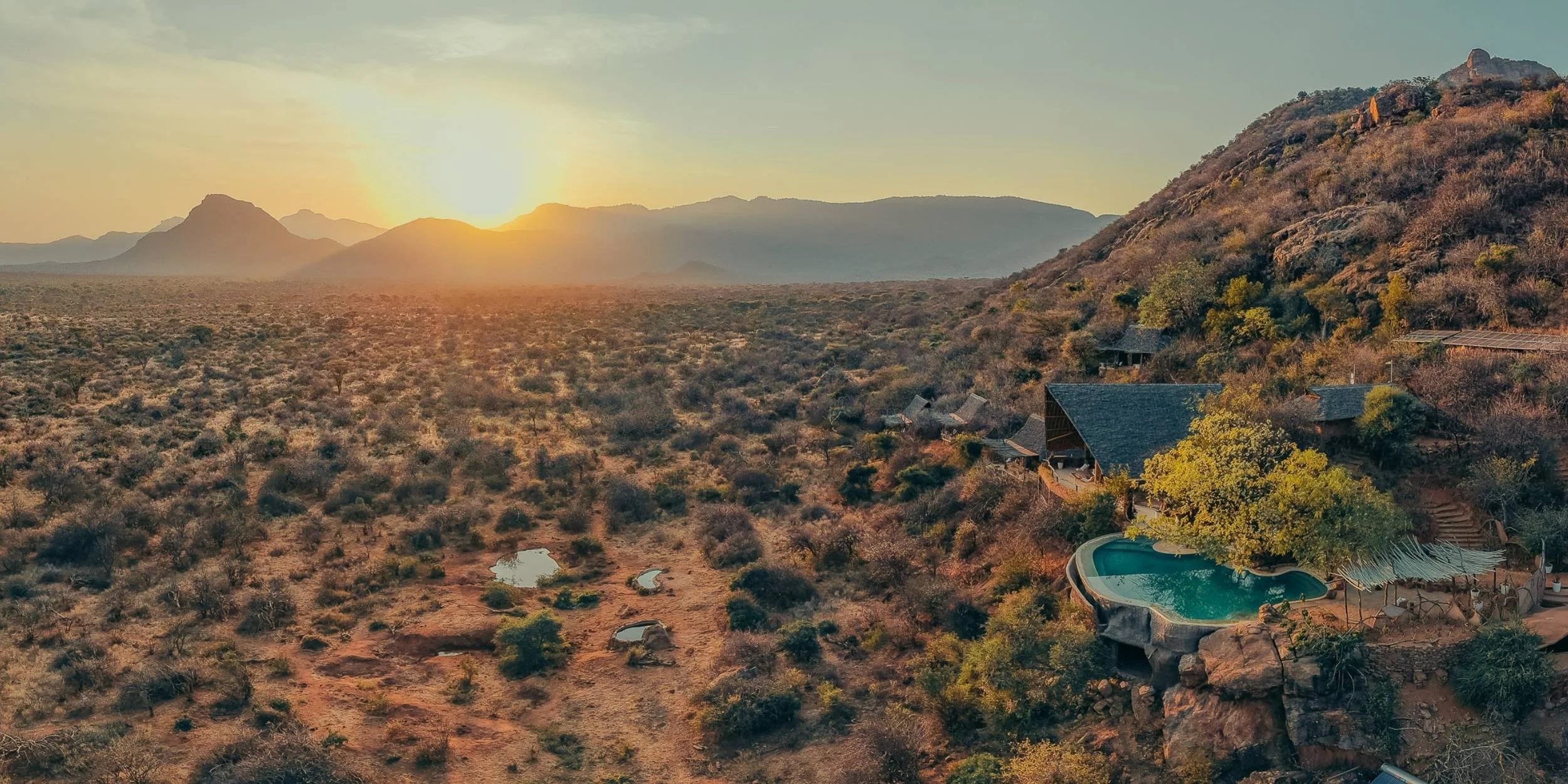 A scenic view of a desert landscape at sunset with mountains in the background, featuring a house with a swimming pool and surrounding vegetation.
