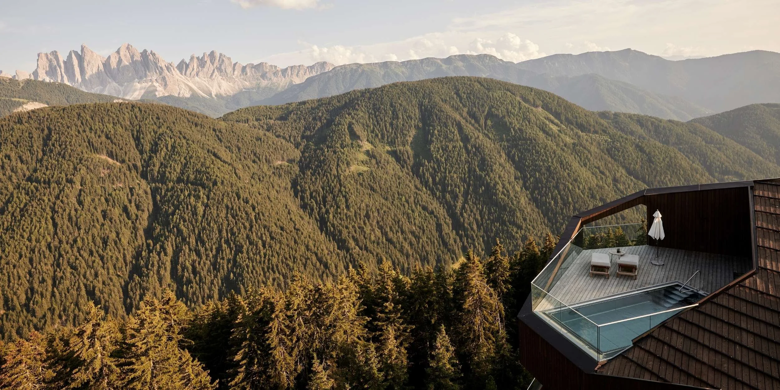 A mountain landscape with green, forested hills and distant rocky mountain peaks under a partly cloudy sky. In the foreground, there is a modern balcony with a small swimming pool, two lounge chairs, a closed umbrella, and glass railing.