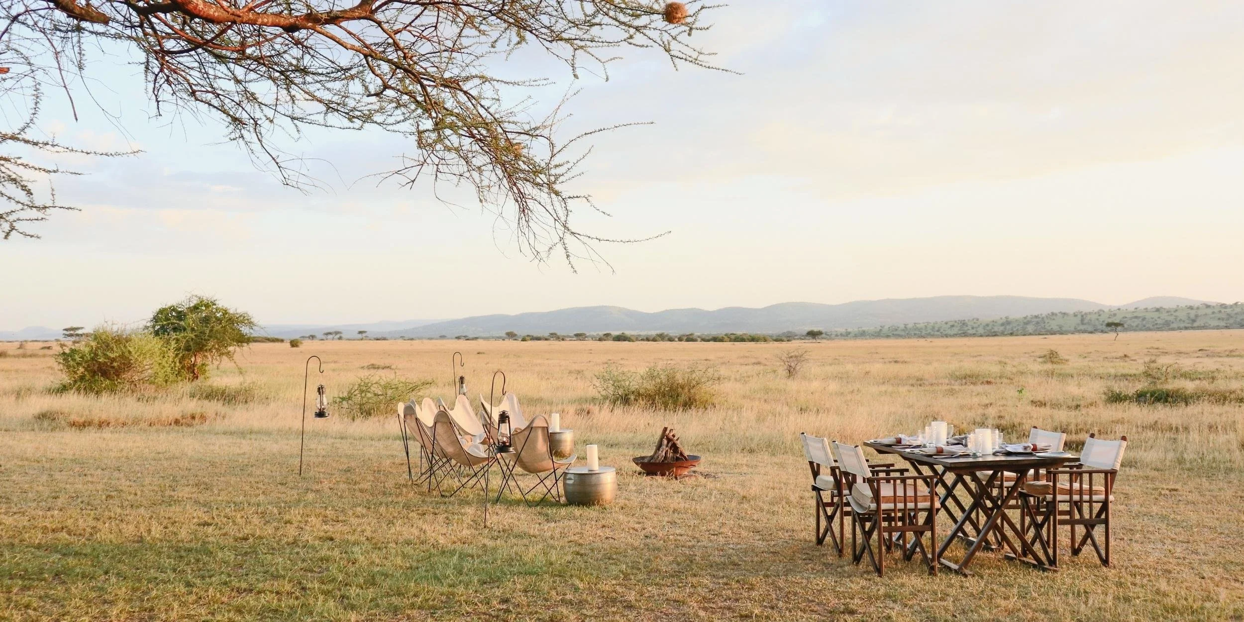An outdoor dining setup in a savannah, with a large wooden table and chairs, plates, glasses, and candles. Nearby, there are camping chairs, lanterns, and a small fire pit, with expansive grassland and distant mountains in the background.