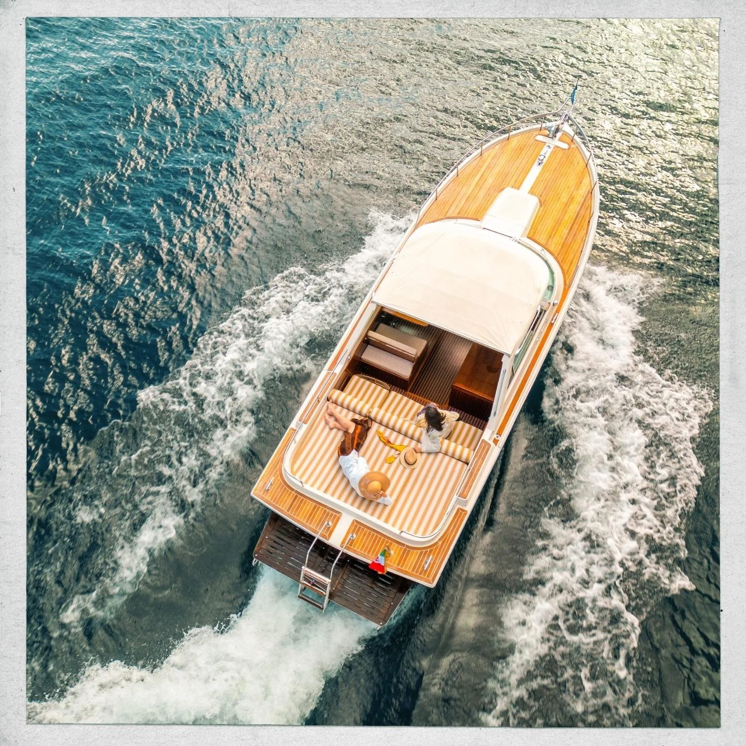 An aerial view of a boat sailing on the water with two women relaxing on the deck under a canopy, enjoying the sunny day.