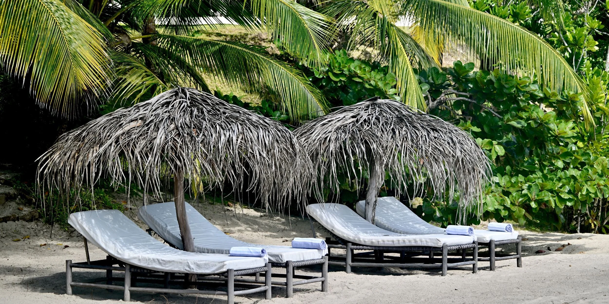 Three lounge chairs with rolled towels under thatched umbrellas on a sandy beach with palm trees and green foliage in the background.