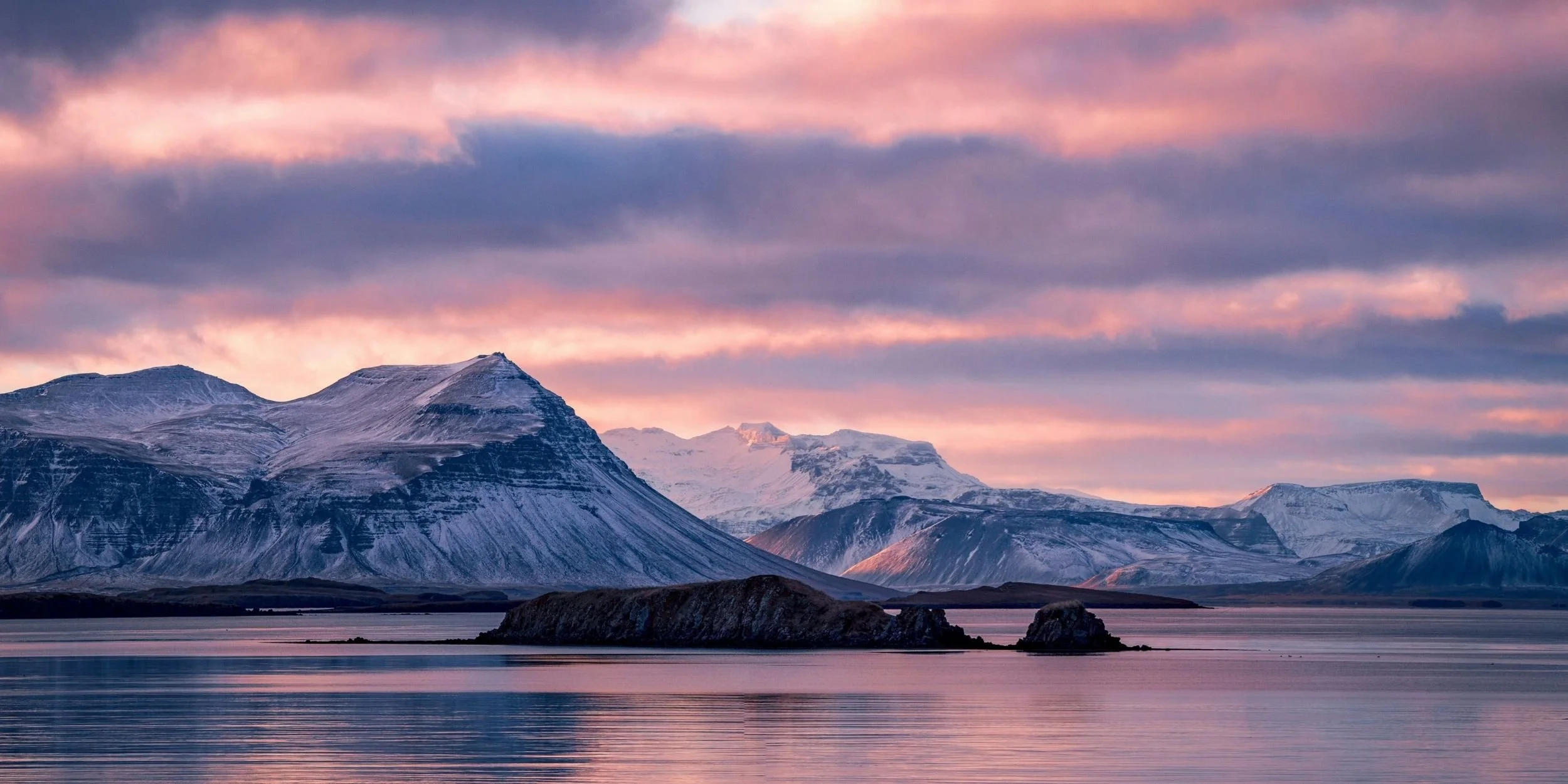 Snow-capped mountains beside a calm body of water with a pink and purple sunset sky.