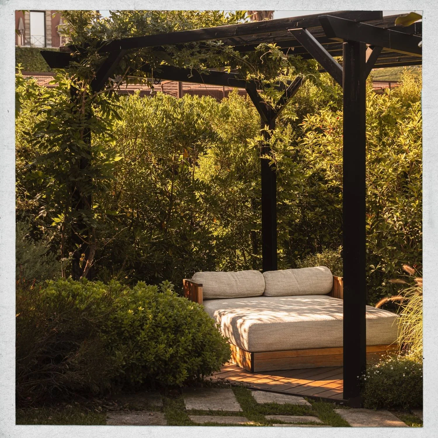 A garden seating area with a cushioned sofa beneath a black pergola surrounded by lush green bushes and trees in sunlight.