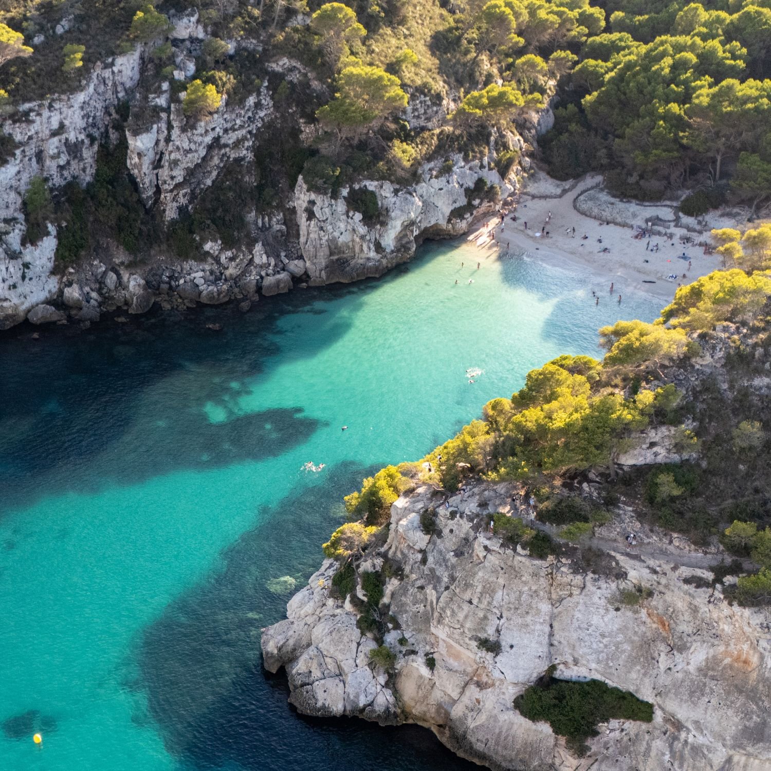 Aerial view of a small beach cove with turquoise water, surrounded by rocky cliffs and green trees, with people relaxing on the sand and swimming in the water.