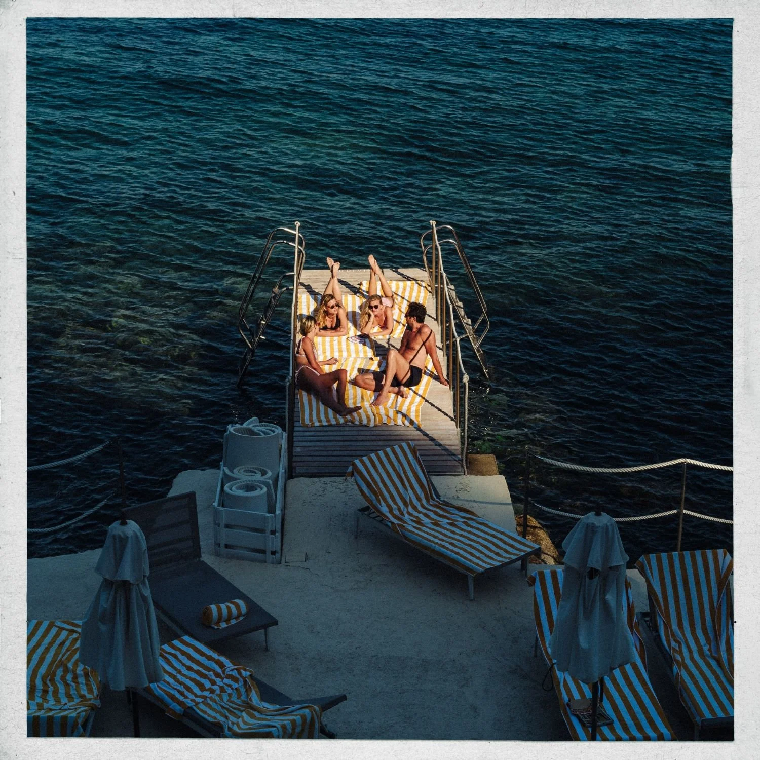 A group of five friends relaxing and chatting on a striped yellow and white lounge bed at a dock over dark blue water, with empty lounge chairs and umbrellas nearby.