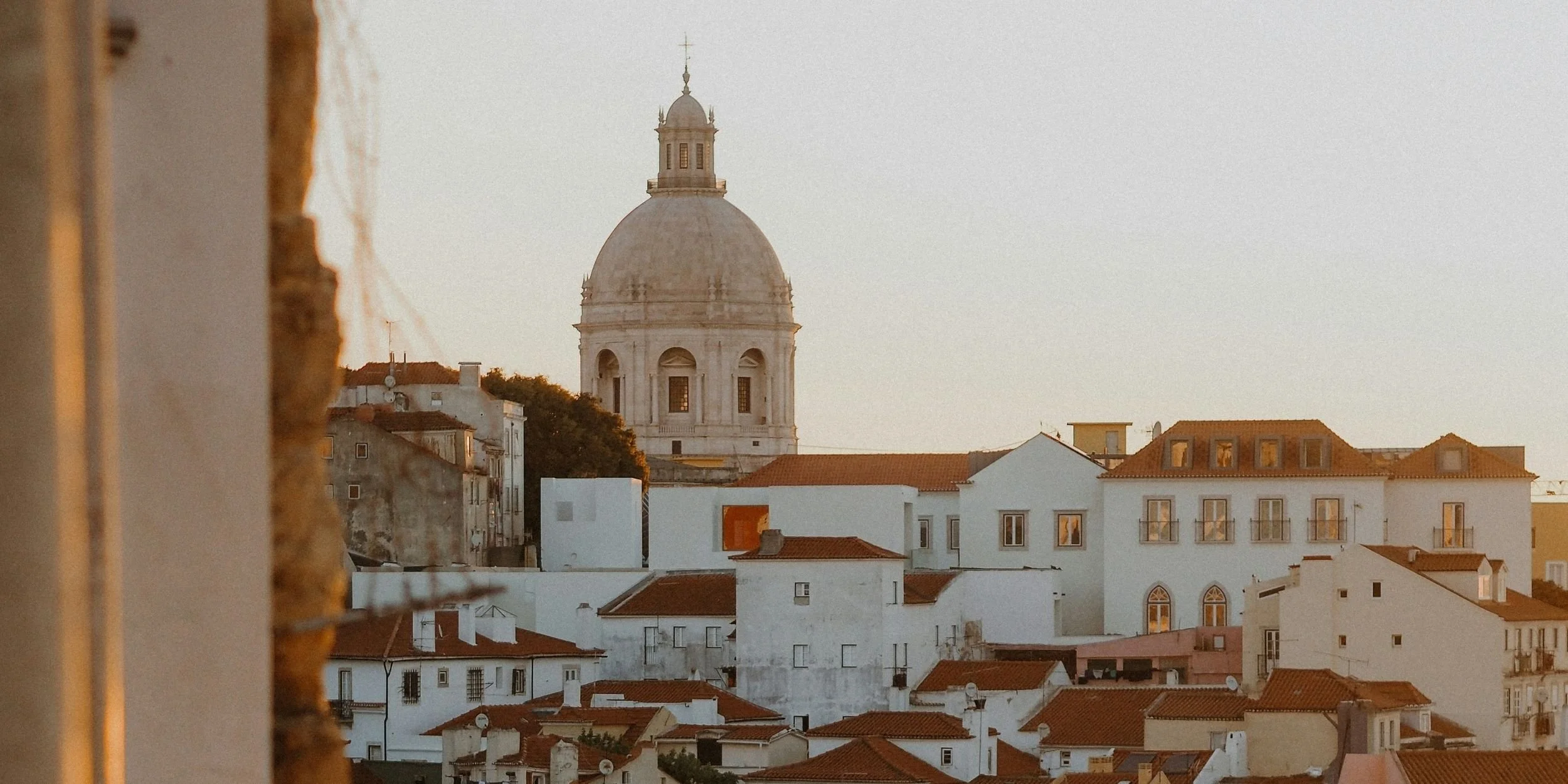 Cityscape with white buildings, red-tiled roofs, and a large domed church in the background at sunset.