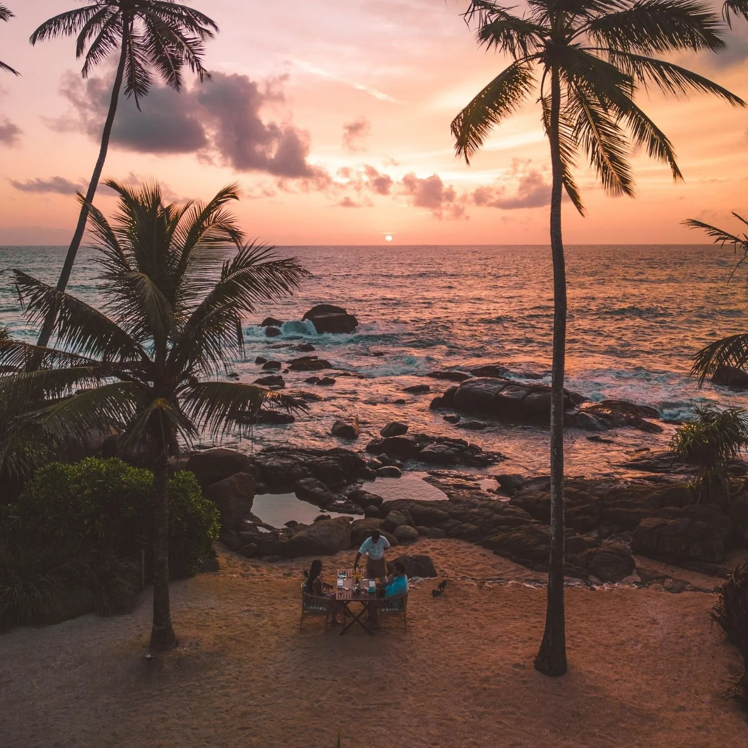 A group of people dining at a table on a sandy beach during sunset with palm trees, rocks, and the ocean in the background.