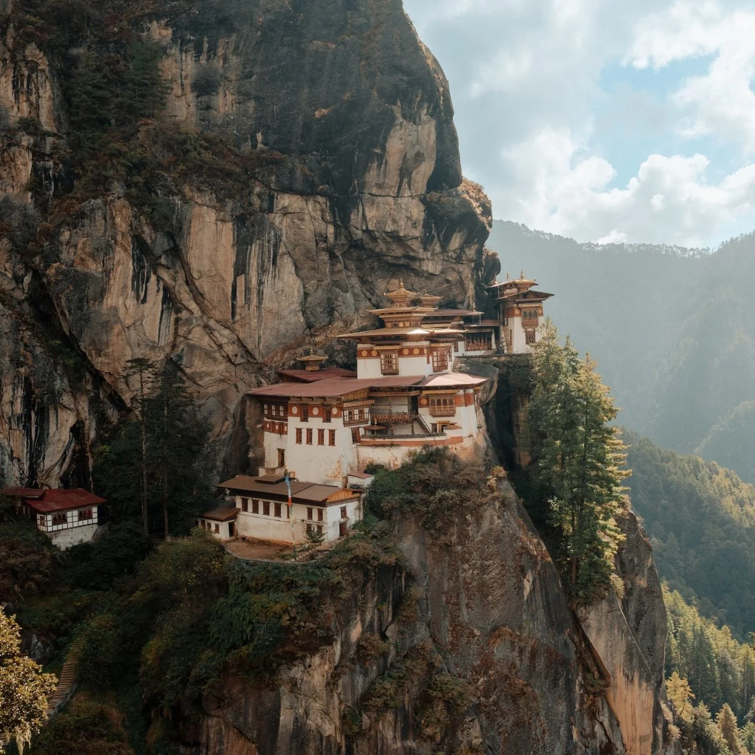 A cliffside monastery built into the steep rocky face of a mountain, with multiple traditional buildings featuring ornate roofs and wooden details, surrounded by trees and forested landscape under a partly cloudy sky.