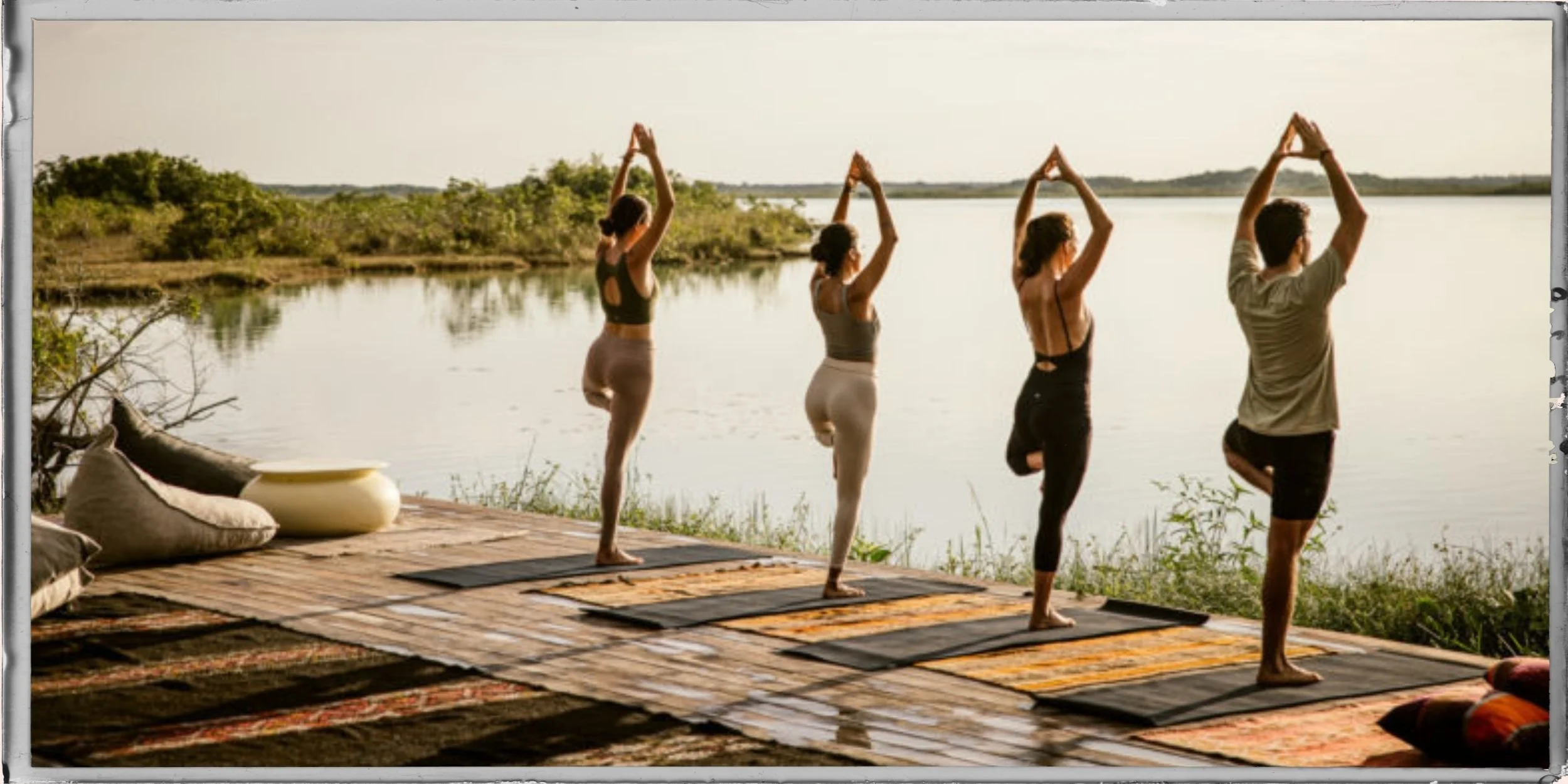 Four people practicing yoga on mats outdoors by a river, performing tree pose with hands together overhead, against a scenic backdrop of water and greenery.