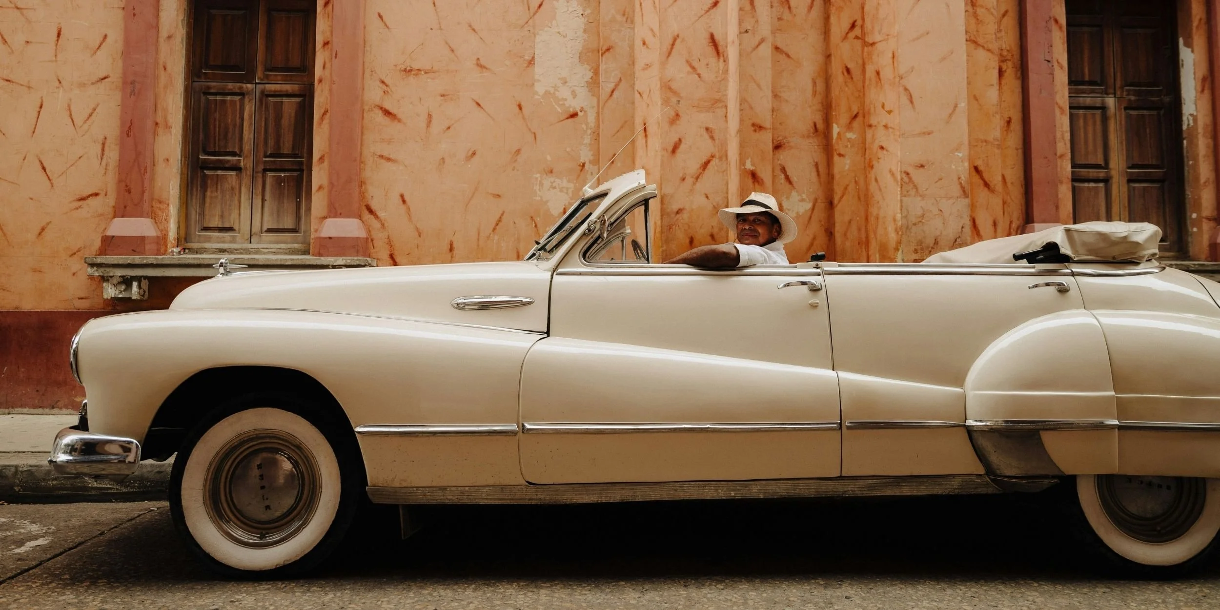 A vintage cream-colored convertible car parked in front of a weathered peach-colored wall with wooden windows. A man wearing a white hat and shirt is sitting in the driver's seat, looking towards the camera.