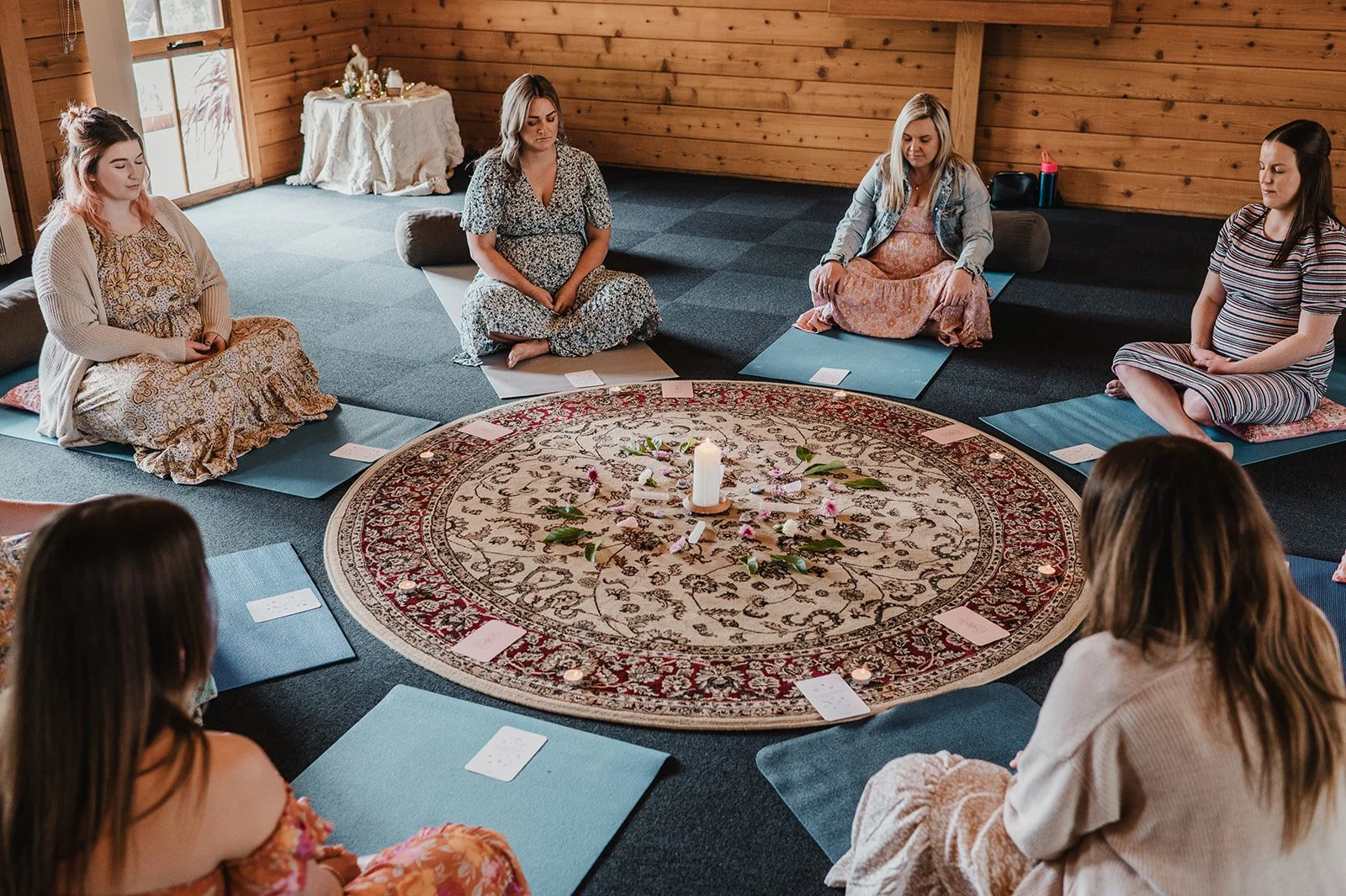 A group of seven women sitting cross-legged on yoga mats arranged in a circle around a decorated circular rug with a candle and flowers in the center, inside a wooden room.