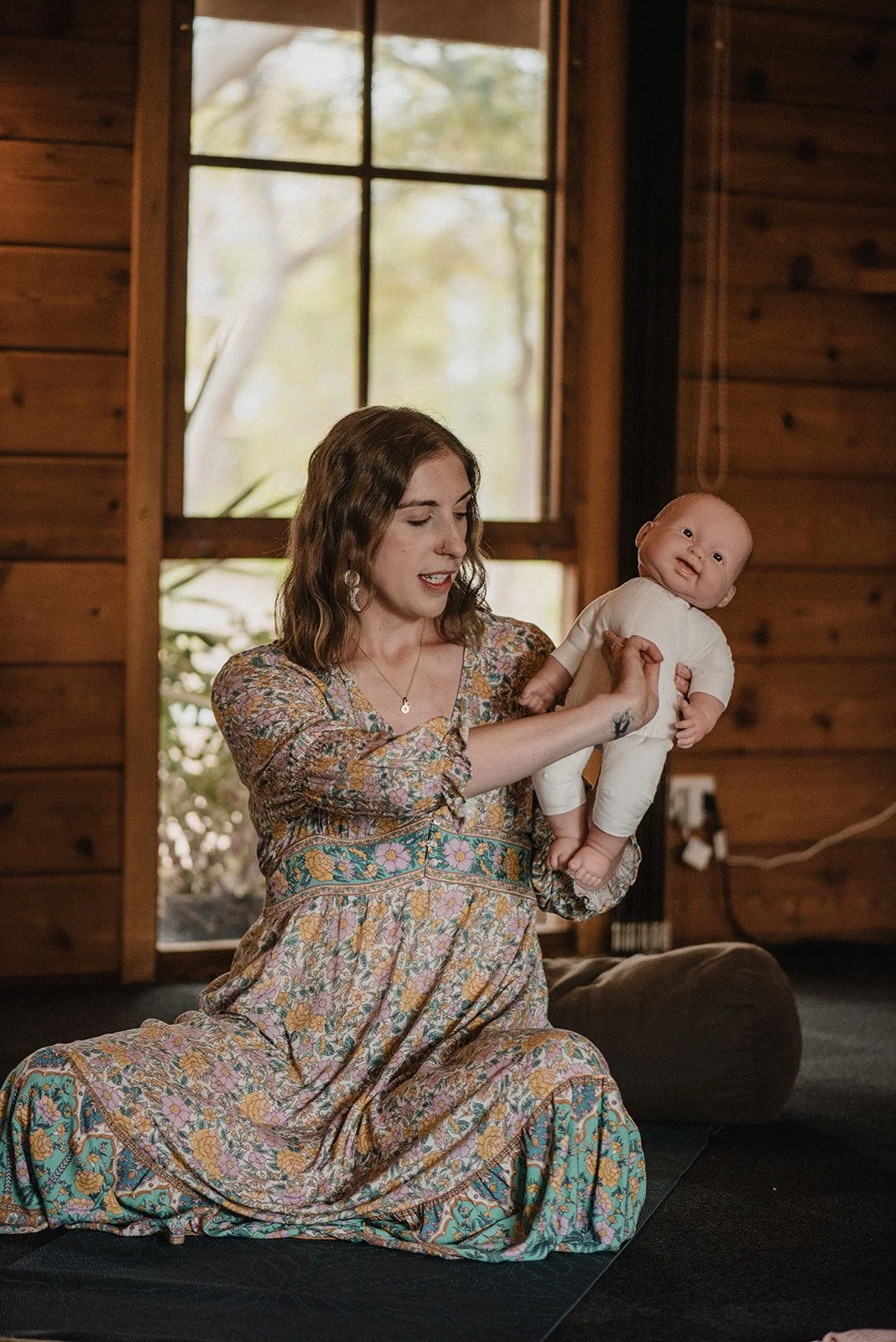 A woman with wavy brown hair holding a smiling baby in a cozy wooden room with large windows showing greenery outside.