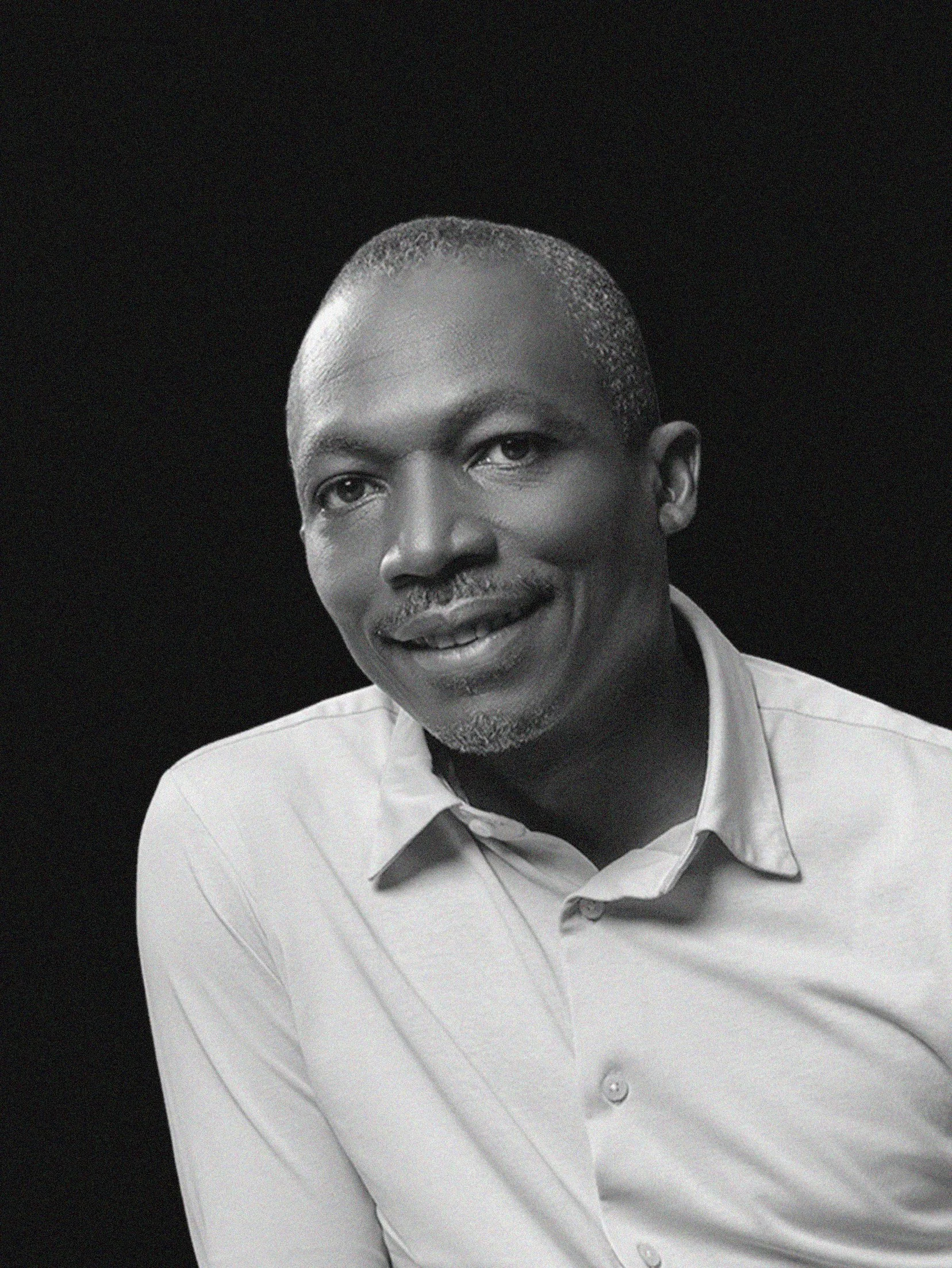 A black-and-white photo of a man sitting at a table, smiling and with his hands clasped. He is wearing a light-colored button-down shirt and appears to be in an indoor setting with a dark wooden wall behind him.