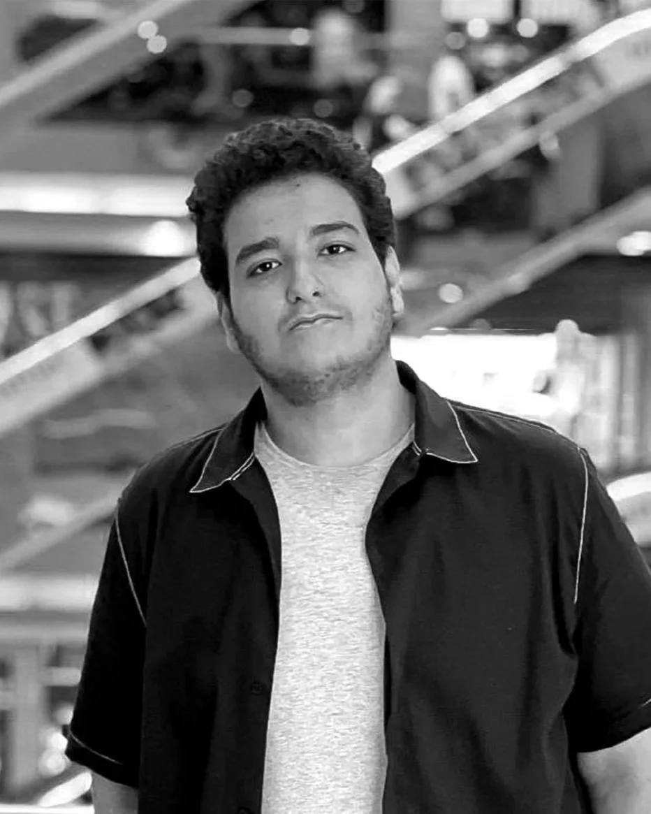 Black and white photo of a young man with curly hair, wearing a casual jacket over a t-shirt, standing in an indoor setting with blurred stairs and people in the background.