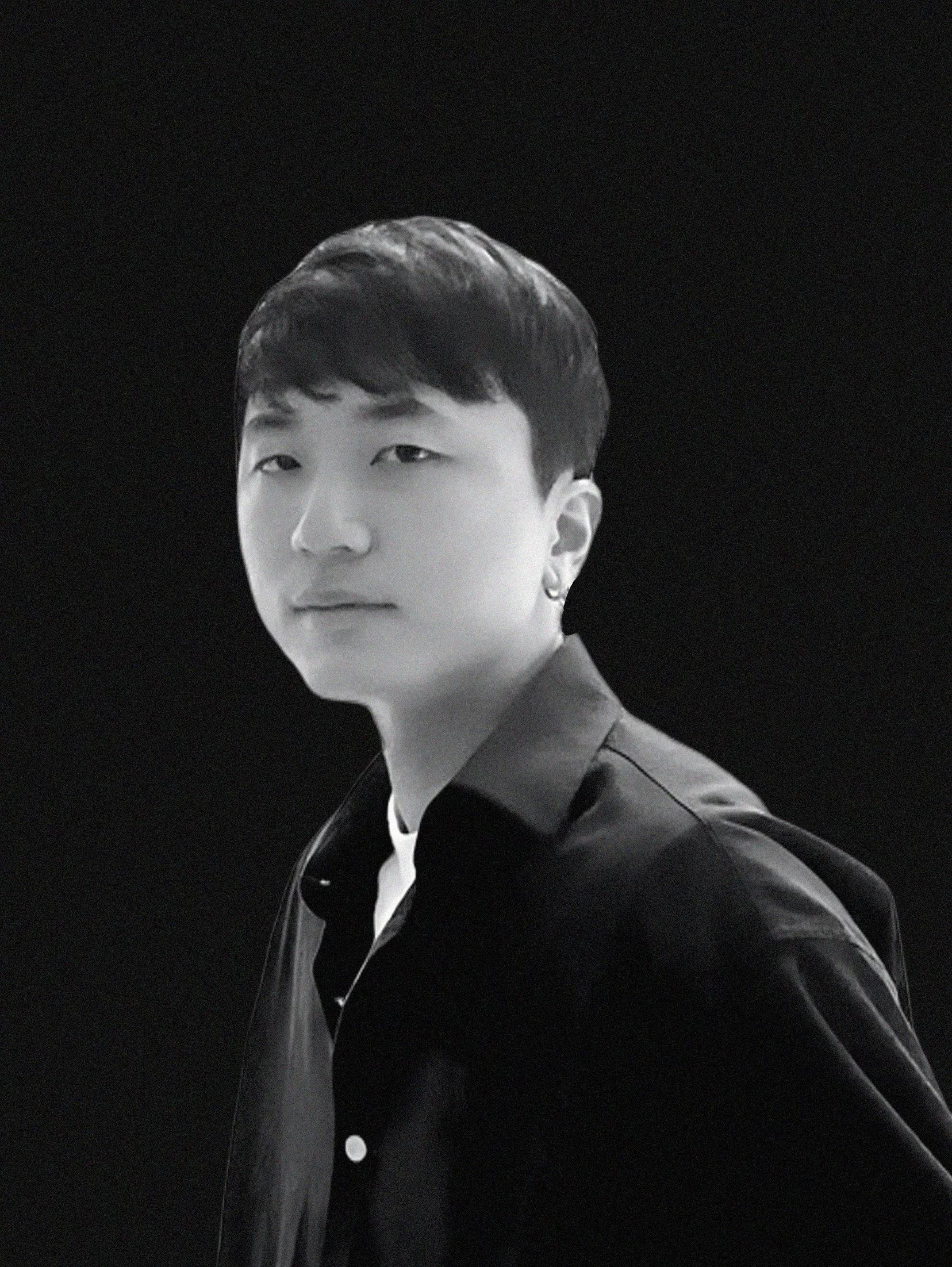 Black and white photo of a young man with dark hair and earrings in an indoor space with large windows, with people in the background.