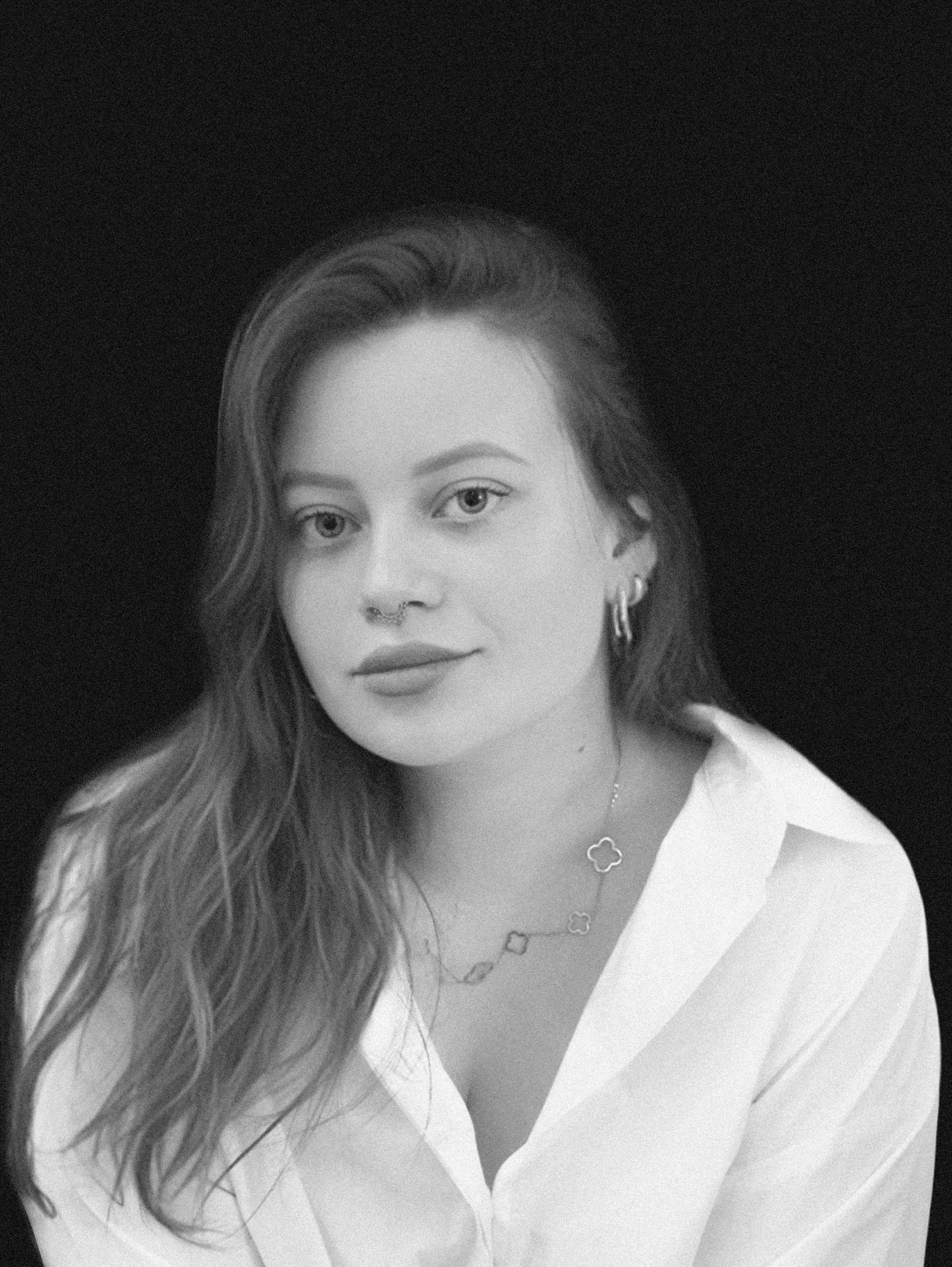 Black and white portrait of a woman with long wavy hair, wearing a white top and jewelry, looking at the camera against a plain white background.