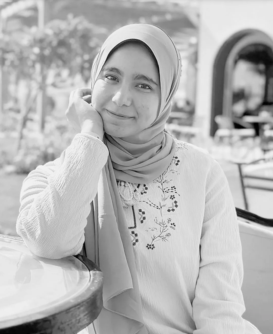 A woman wearing a hijab and a light-colored embroidered top, sitting at a table outdoors, smiling at the camera.